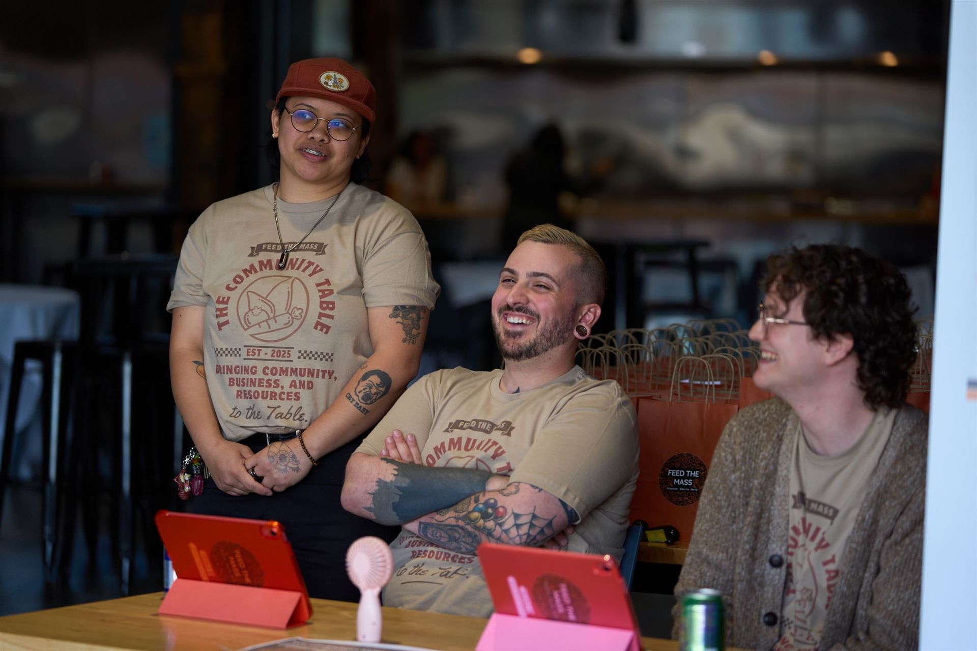 Three people with tattoos and matching shirts smile at a table with iPads in a cafe.
