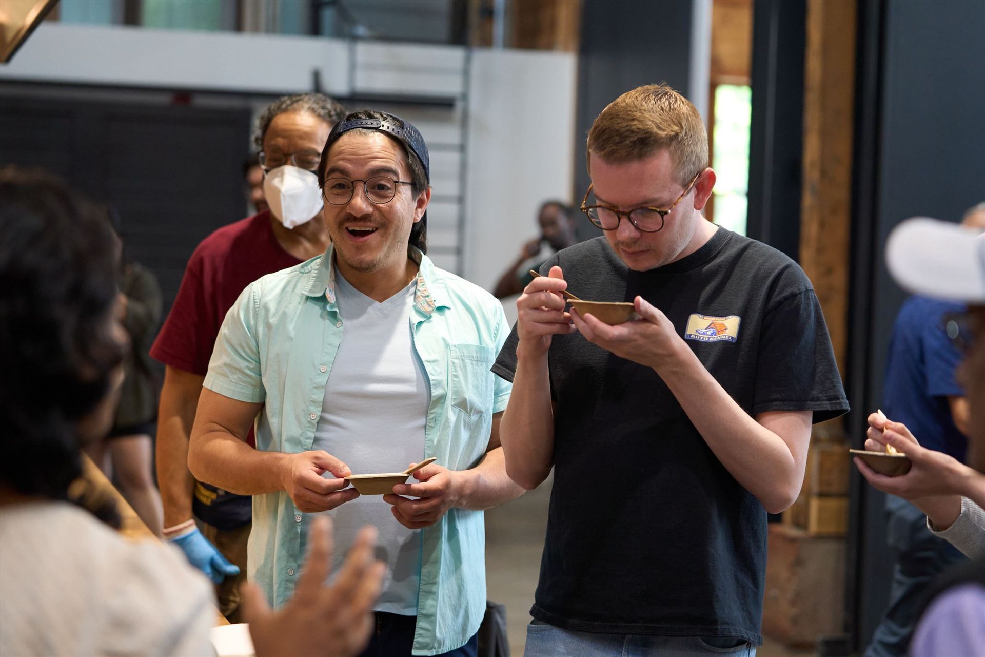 Two men tasting from small bowls, possibly at a food event. People are around them indoors.
