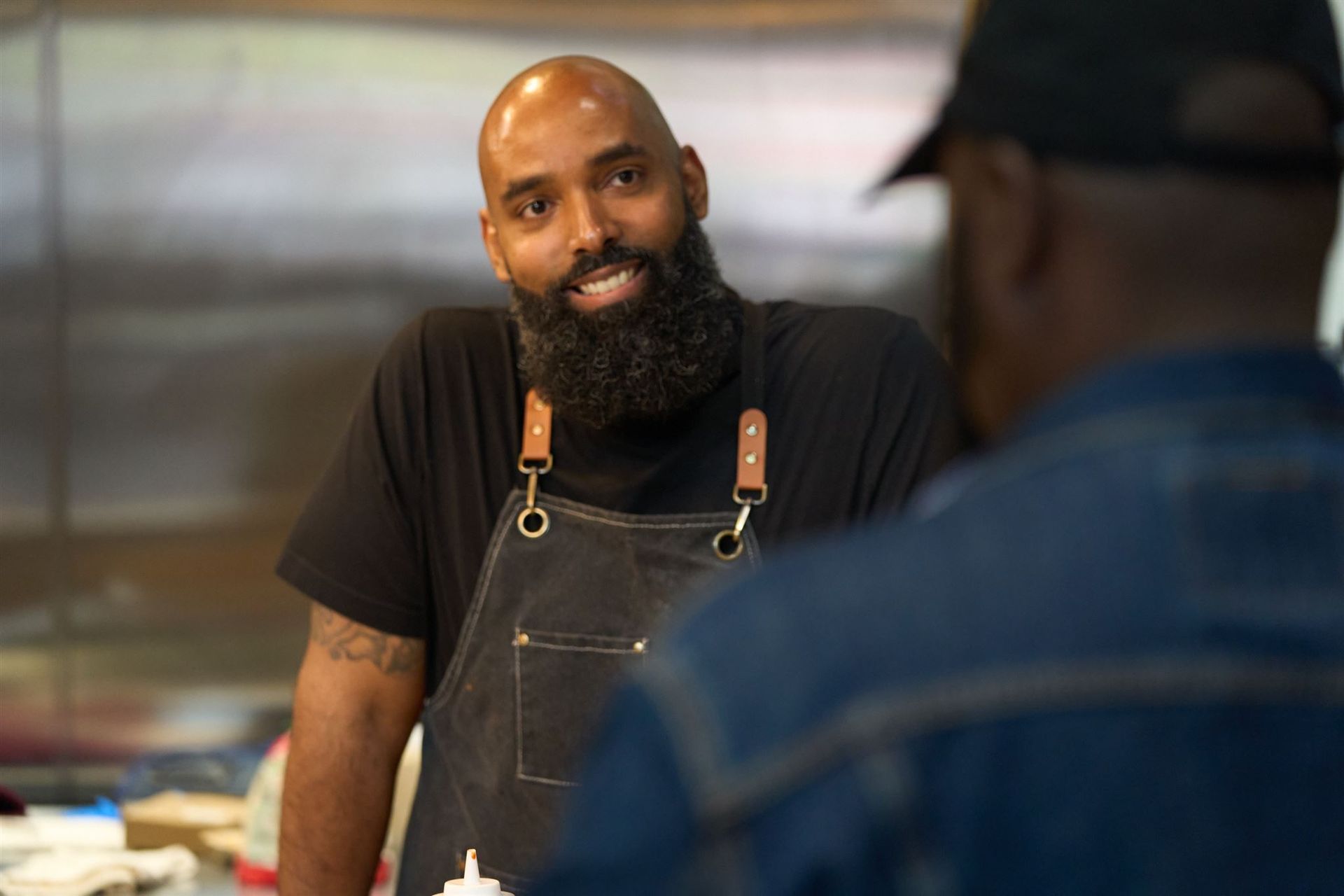 Chef with a long beard smiles, talking to a customer in a kitchen.