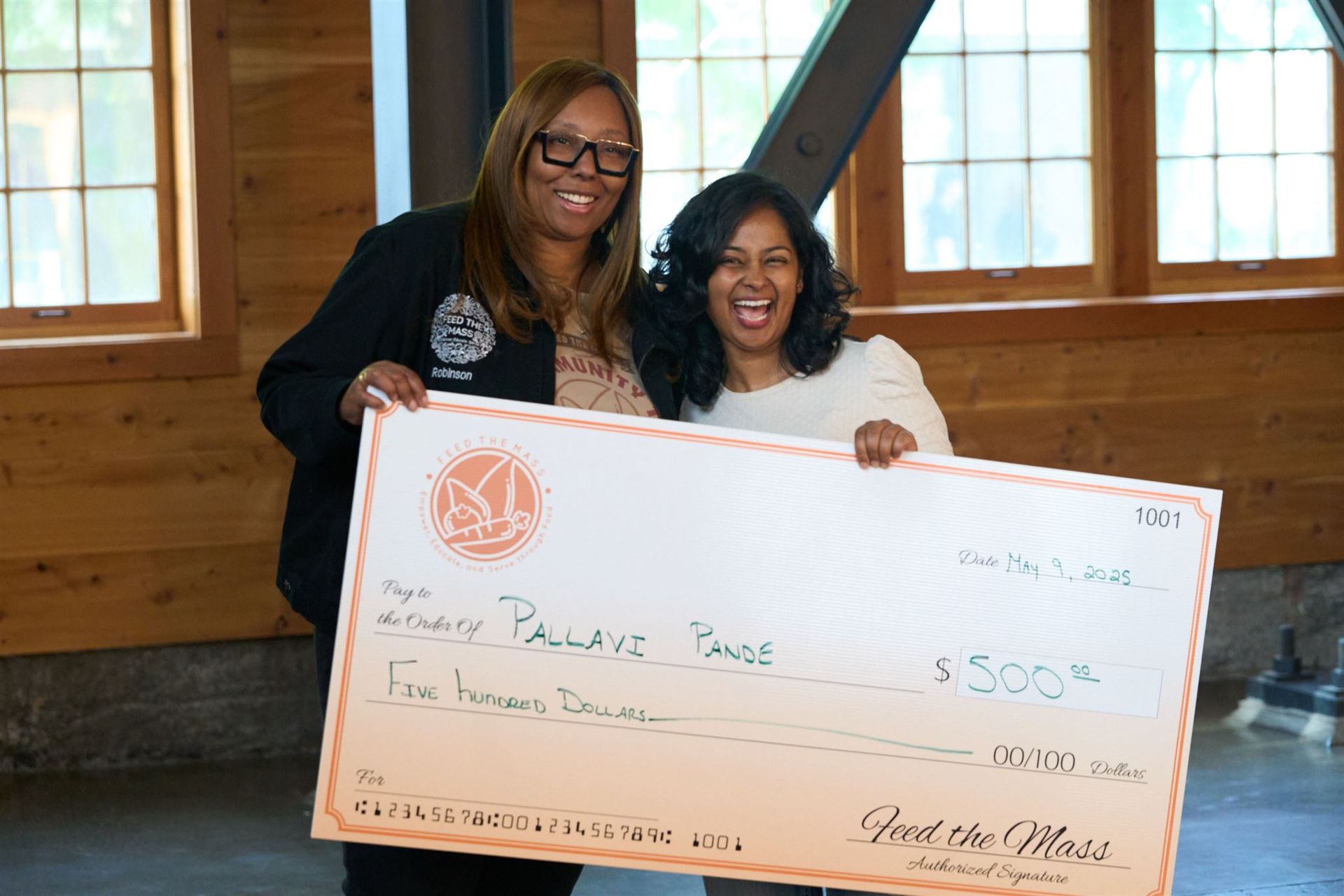 Two women smiling, holding a large check for $500. Light-filled room with windows.