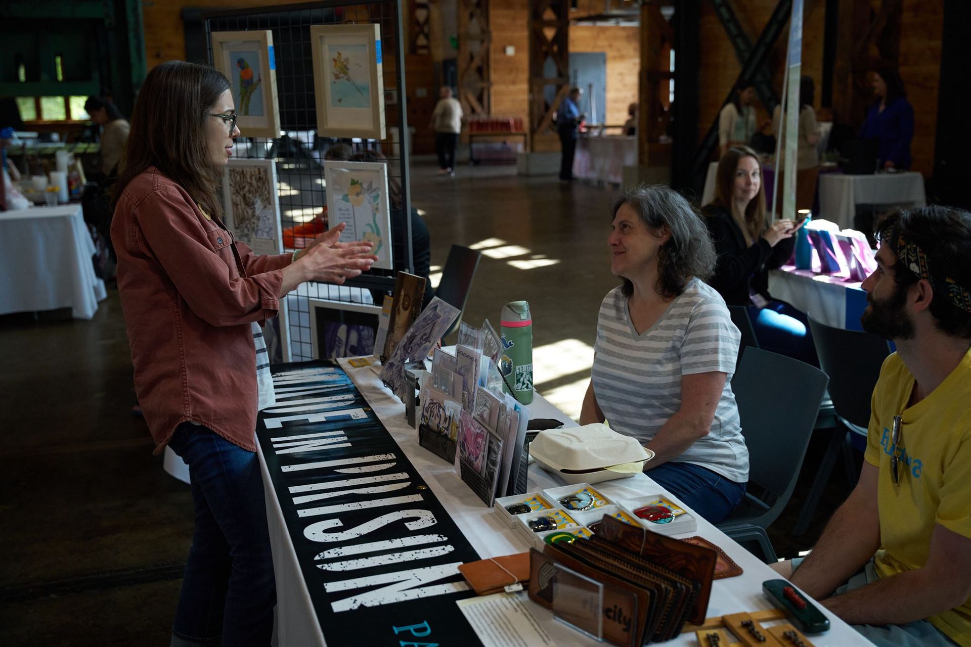 Art fair: Woman explains art to seated customers. Indoor setting with artwork on display.