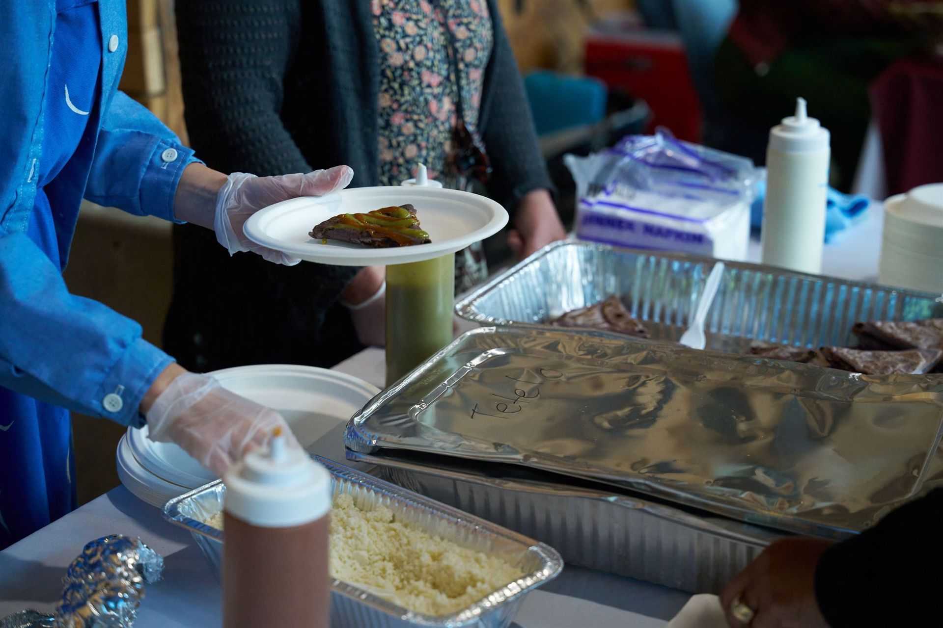 Person serving food from a buffet. Includes a plate with food, containers, and serving utensils.
