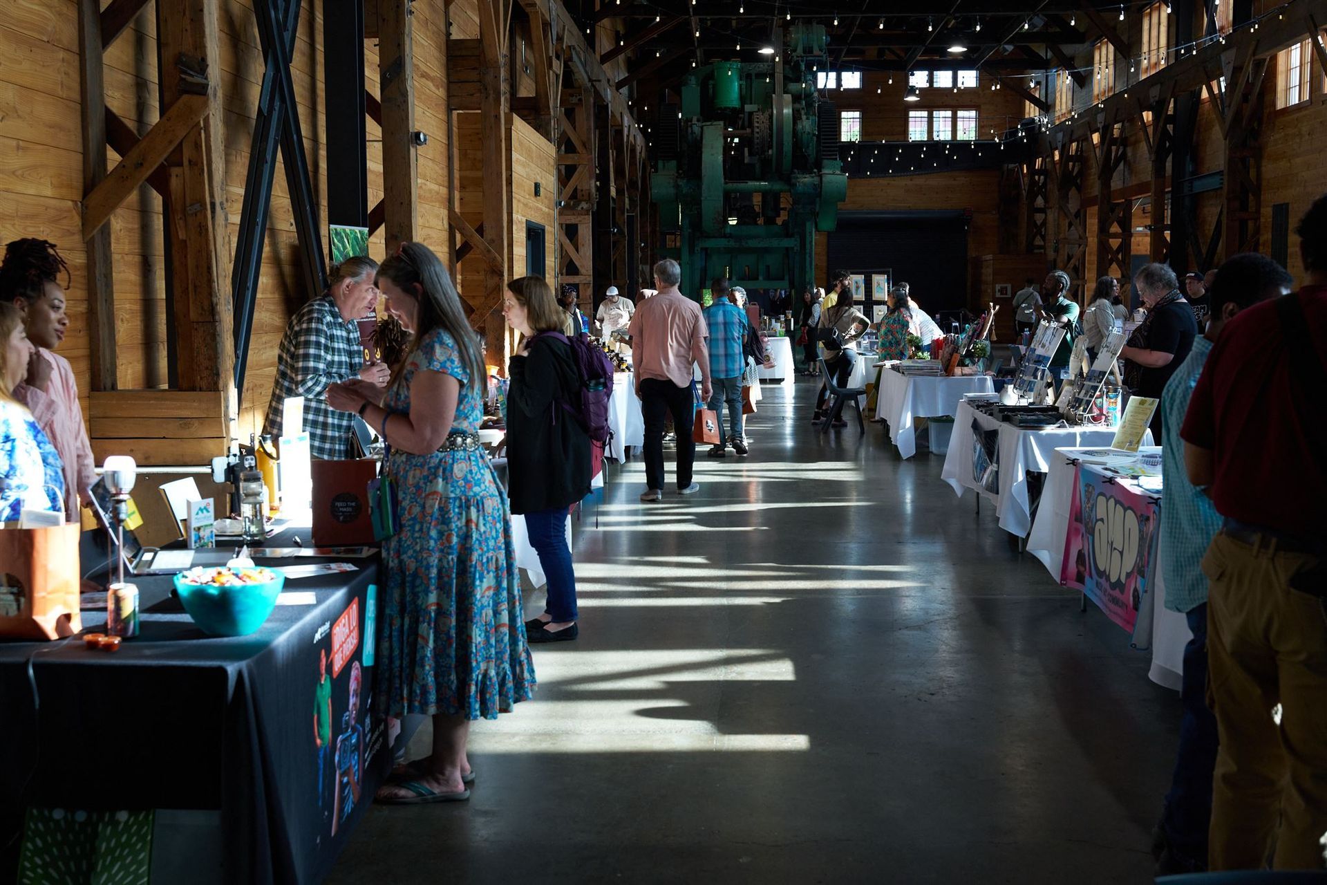 People browsing vendor tables at an indoor market. Sunlight streams through windows.