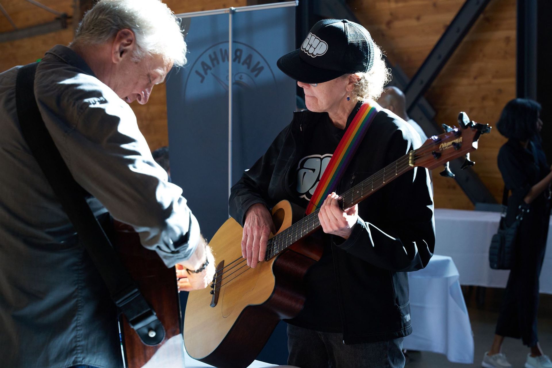 Two musicians playing guitars indoors; one adjusting the other's instrument.