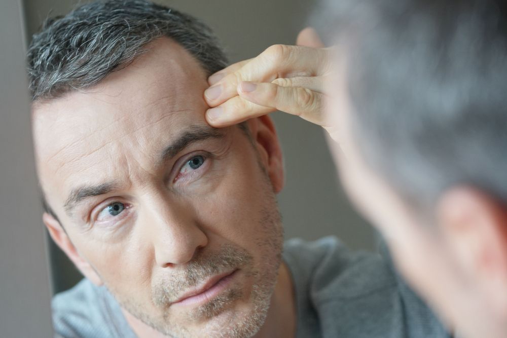 Man Looking in Mirror, Touching Forehead, Examining Wrinkles — Bespoke Institute In Southport, QLD