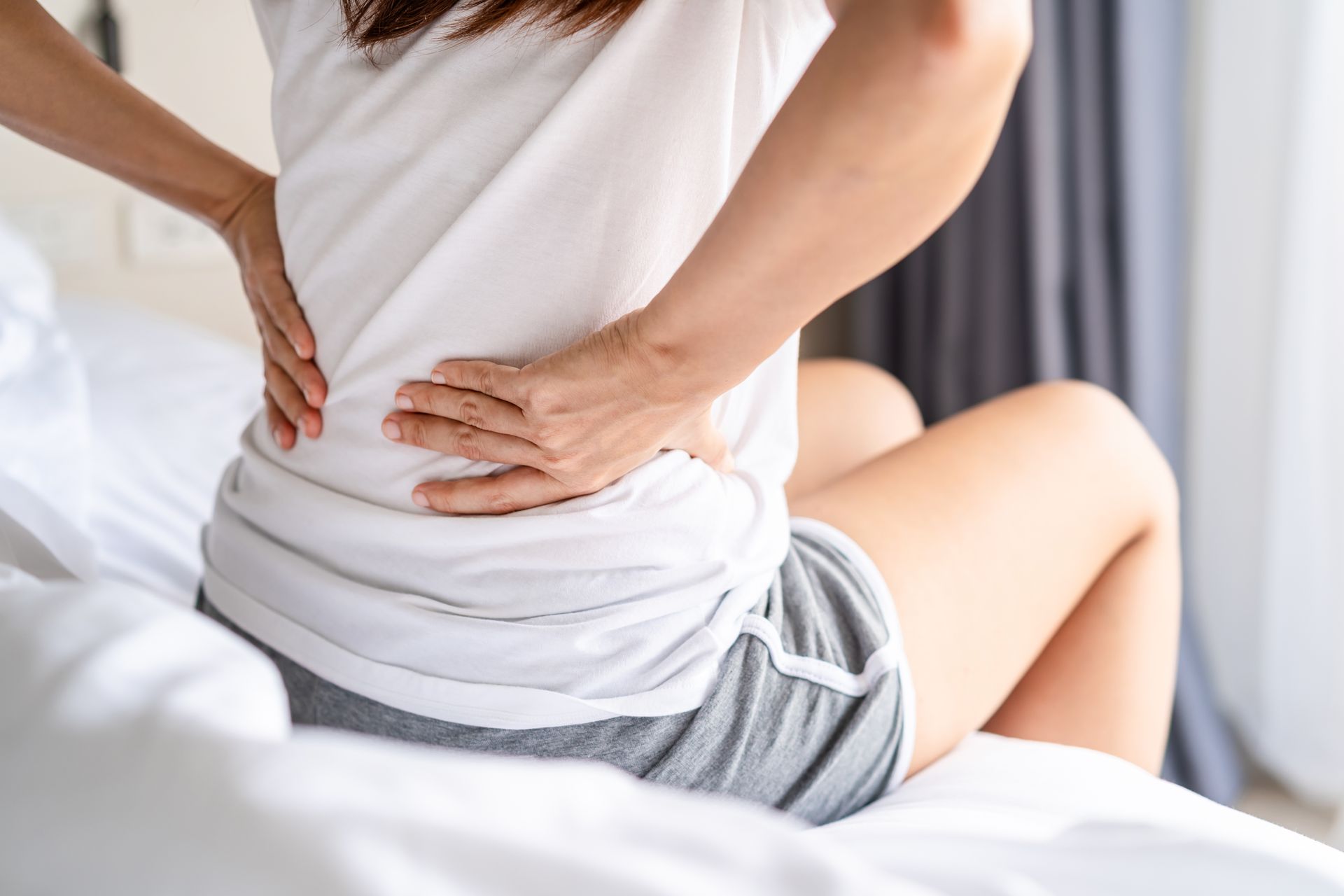 Woman Sitting on Bed, Holding Lower Back in Pain, White Shirt, Grey Shorts — Bespoke Institute In Southport, QLD