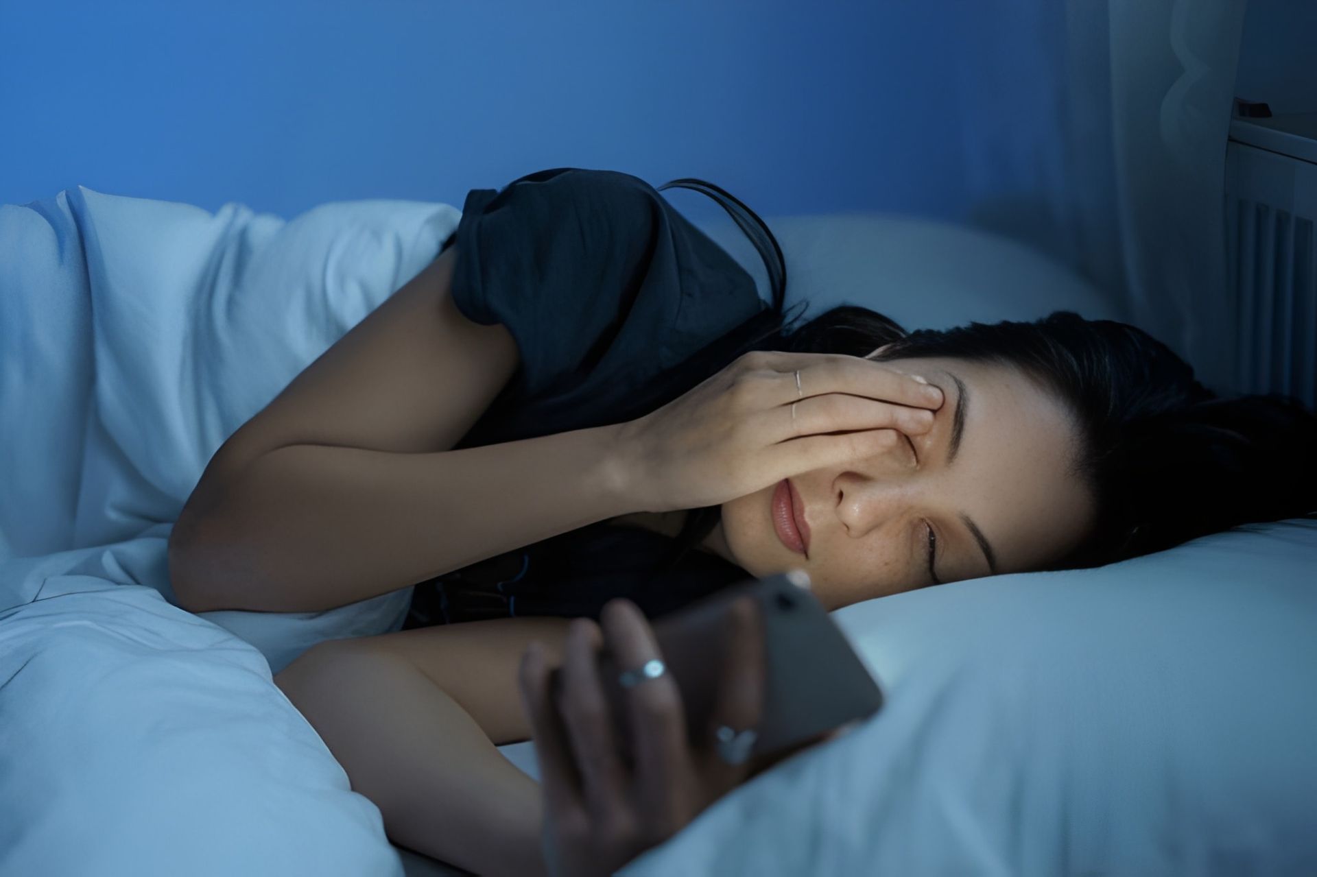 Woman in Bed, Holding Phone, Shielding Eye From Bright Screen — Bespoke Longevity and Aesthetic Medicine In Southport, QLD