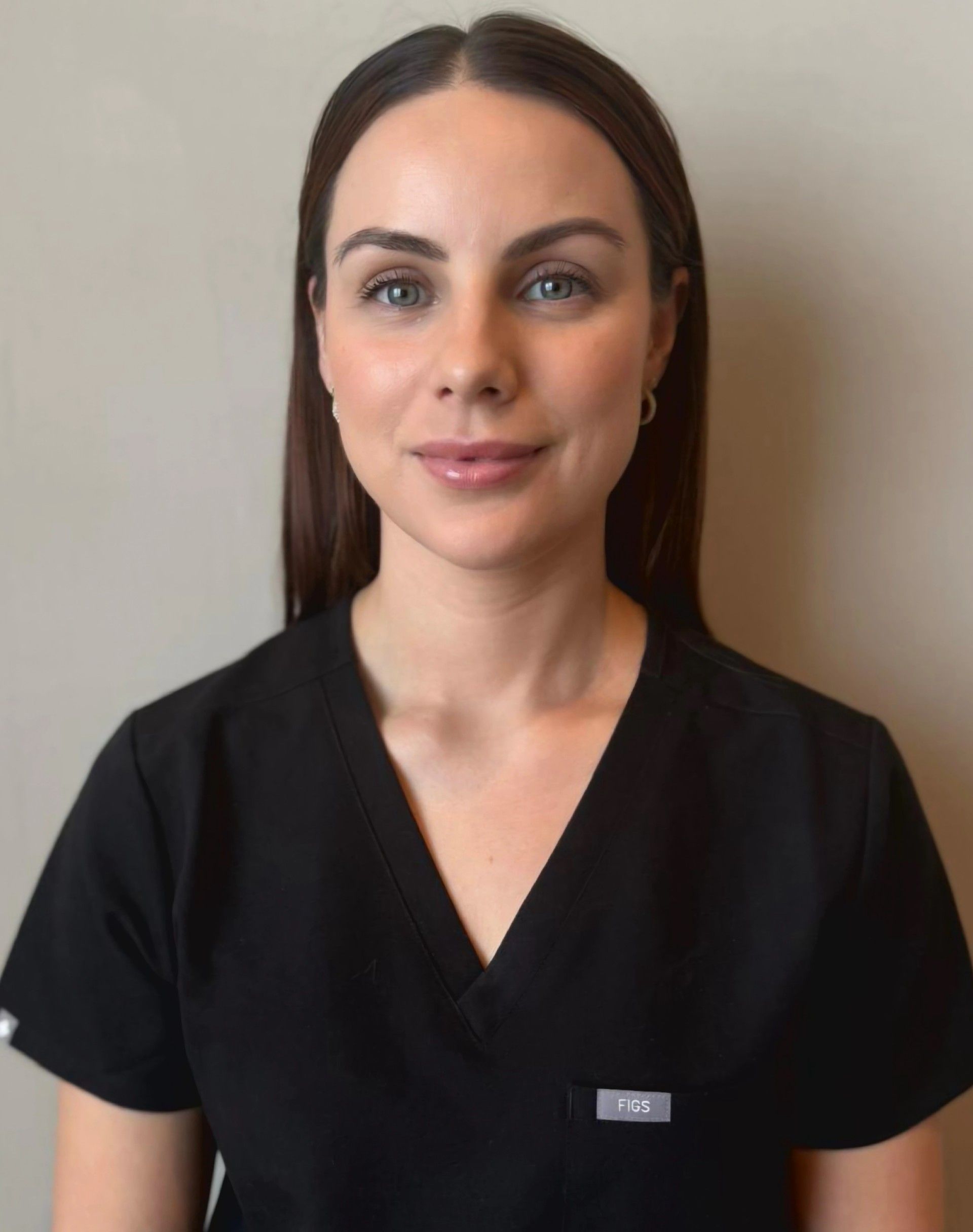 A person with long dark hair, wearing a black v-neck scrub top, posing against a neutral wall.