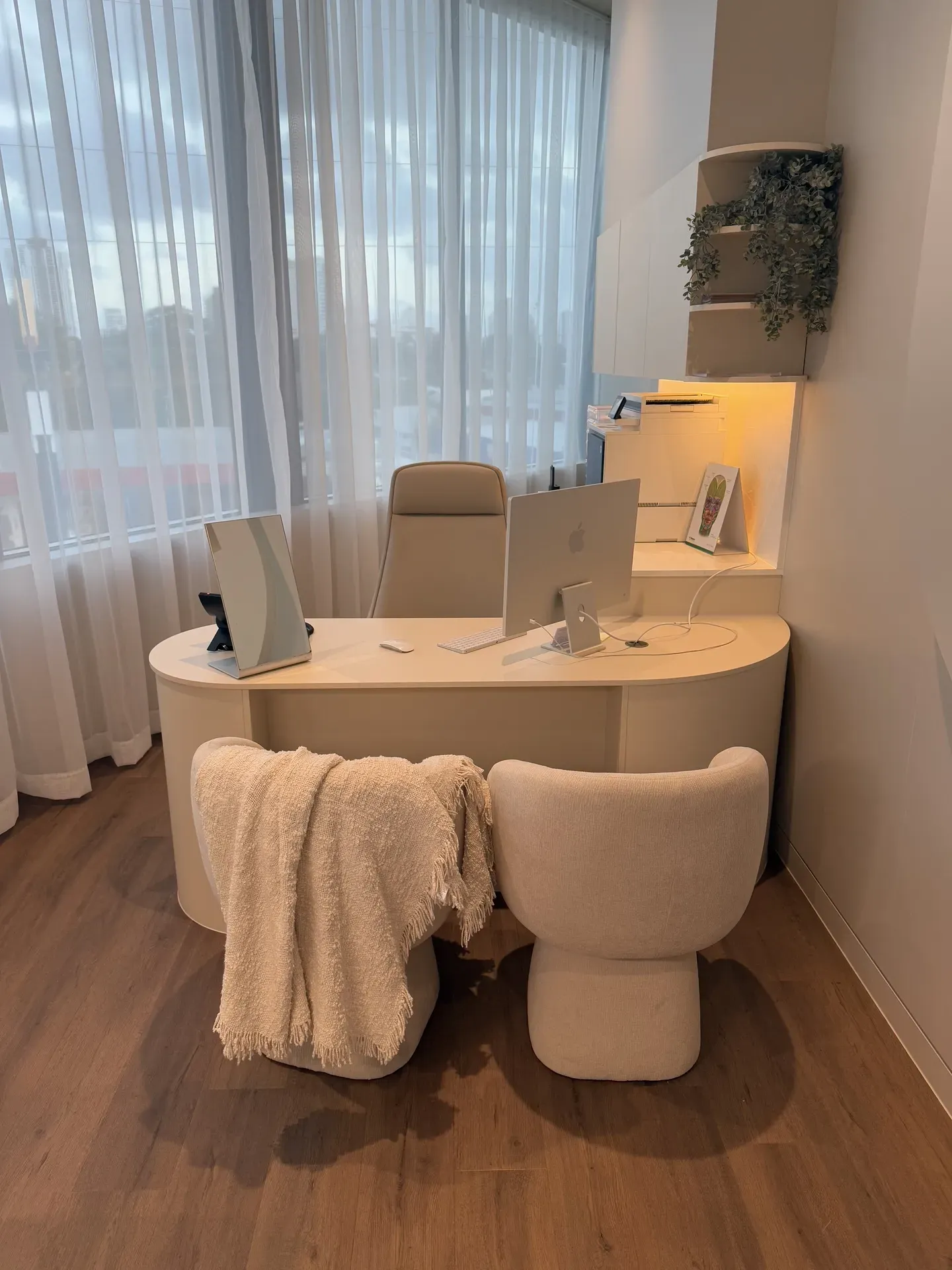 A modern office with a curved desk, two plush chairs, and a window with white curtains.