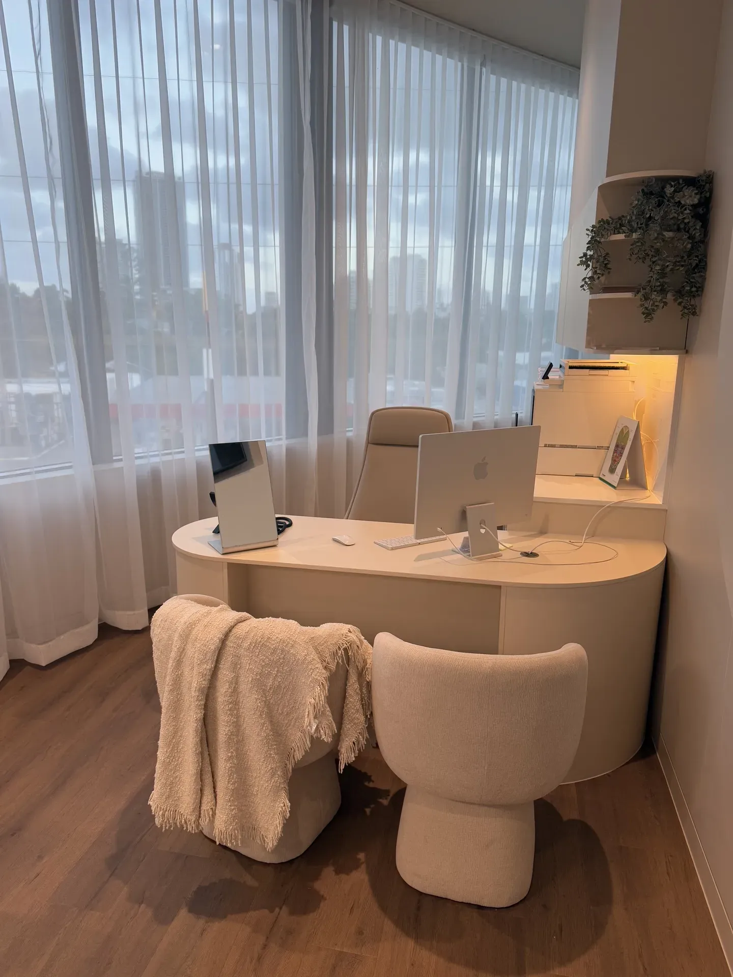 Beige office desk with chair and blanket; window with sheer curtains; computer and monitor.
