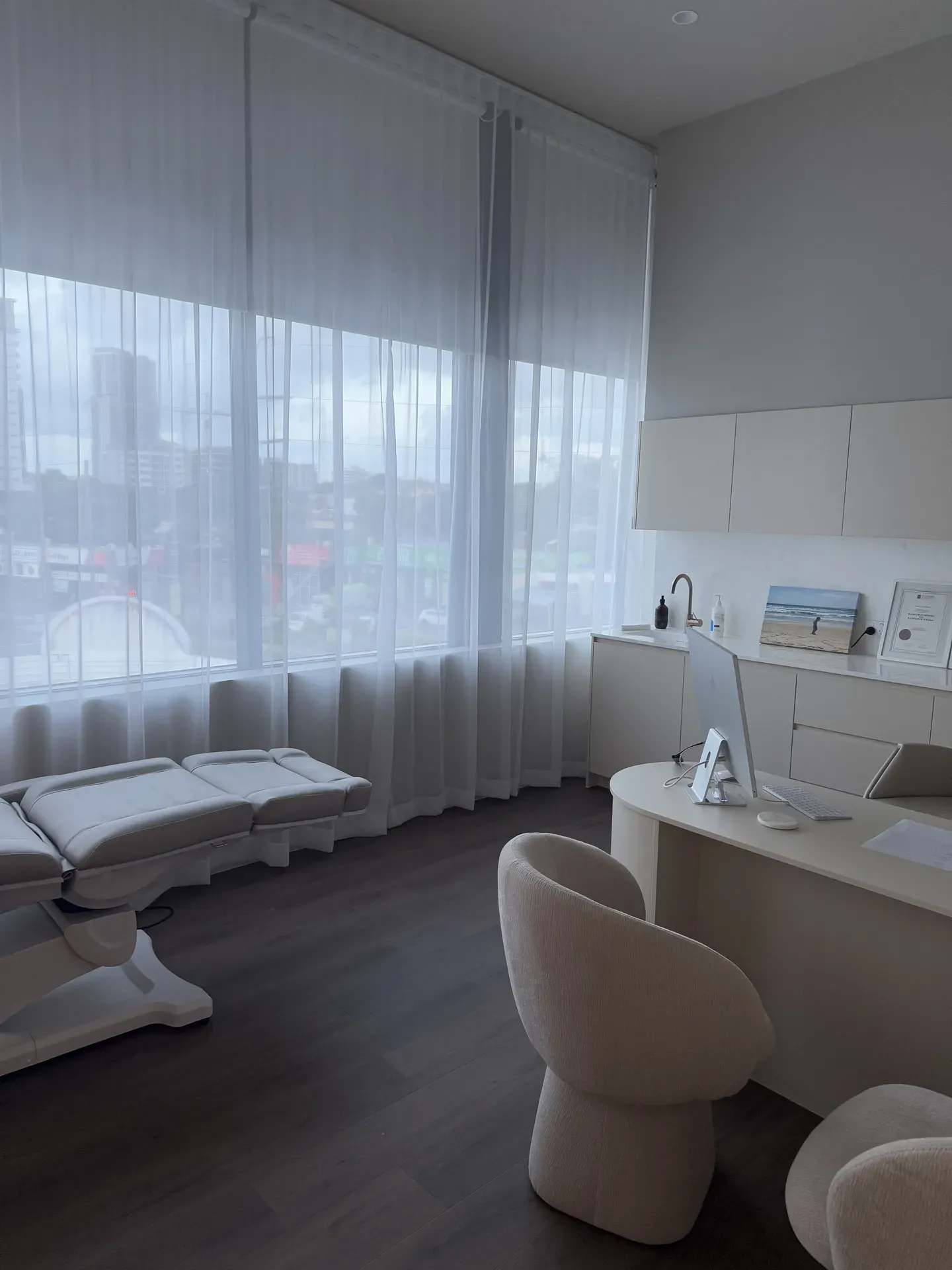 Modern, bright medical office interior with a treatment bed, desk, and large window with sheer curtains.