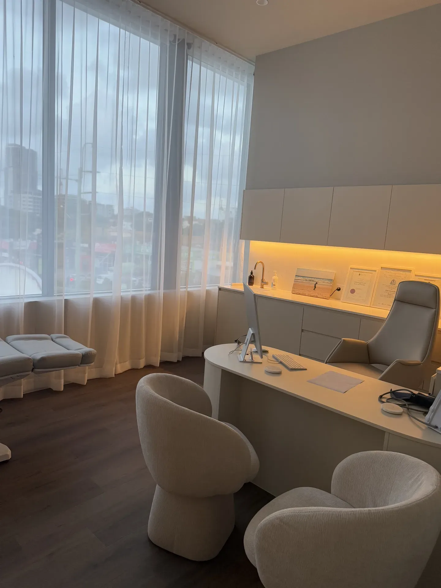 Aesthetician's office. Cream-colored chairs, desk, and treatment bed. Large window with sheer curtains. Wooden floors, bright lighting.