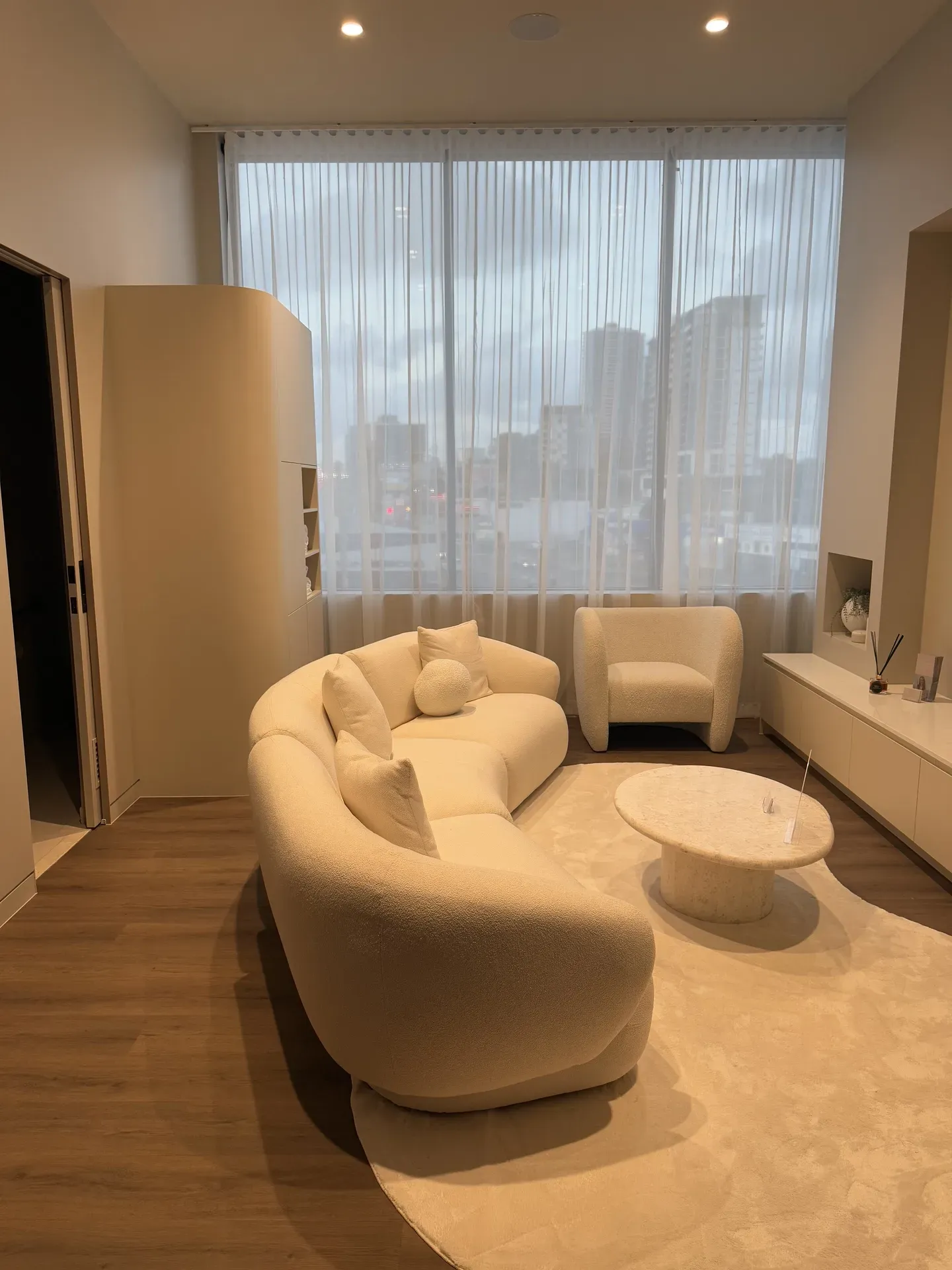 Cozy living room with a cream-colored curved sofa, armchair, and coffee table; large window with sheer curtains.