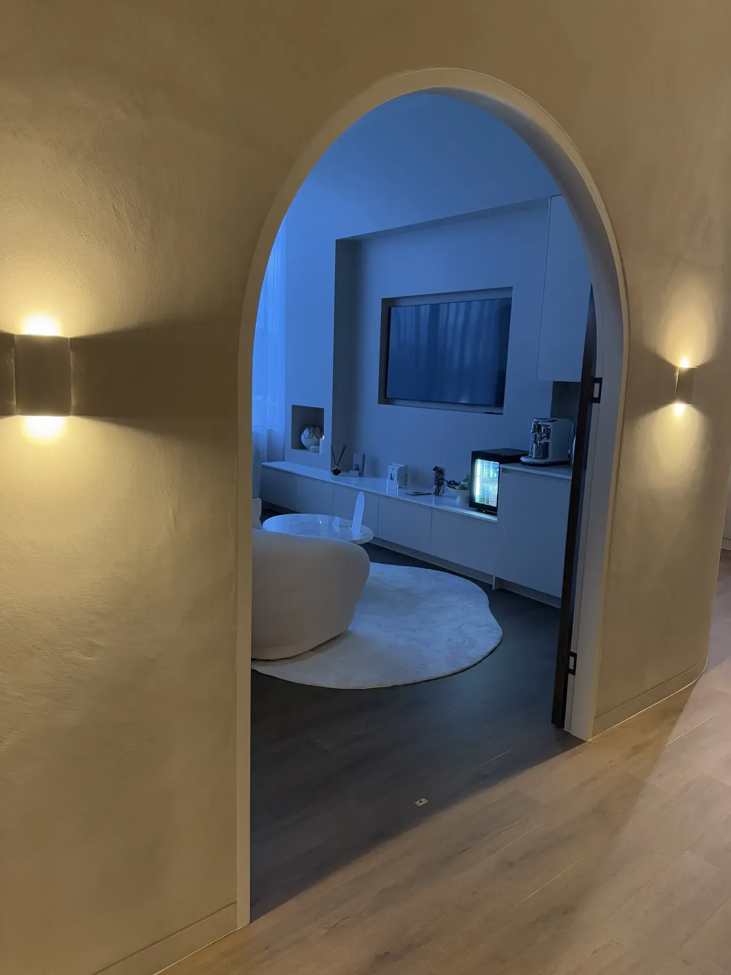 Archway leading to a living room with a TV, white furniture, and a rug. Two wall sconces illuminate the archway.