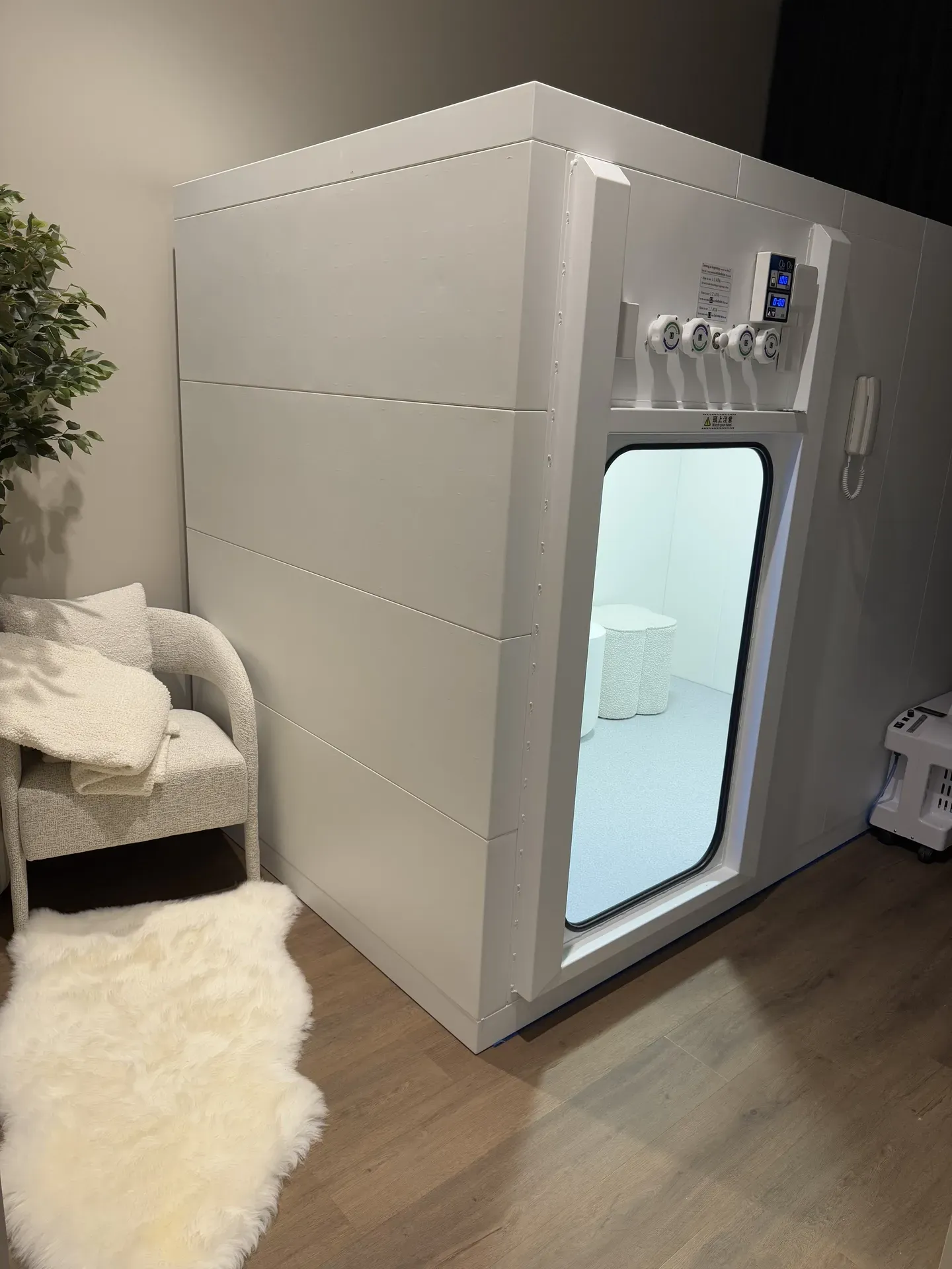 White hyperbaric chamber with a glass door in a corner room, next to a chair and faux fur rug.