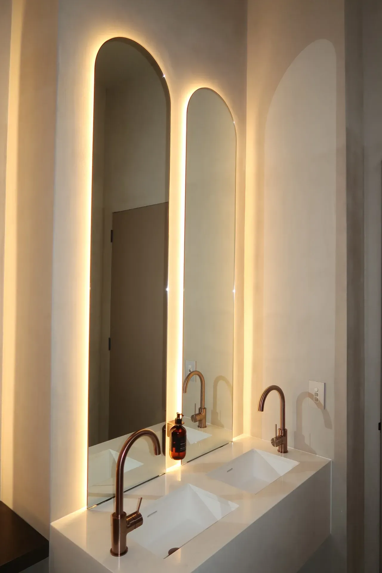 Double sink bathroom with arched mirrors, copper faucets, and warm lighting.