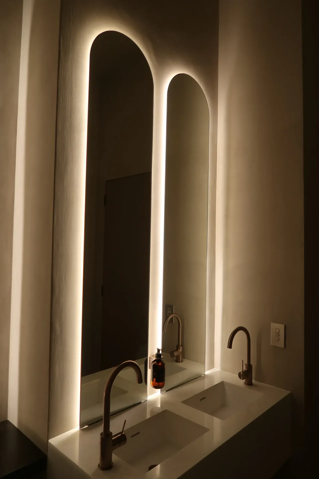 Modern bathroom with arched mirrors, illuminated edges, and two sinks with copper fixtures.