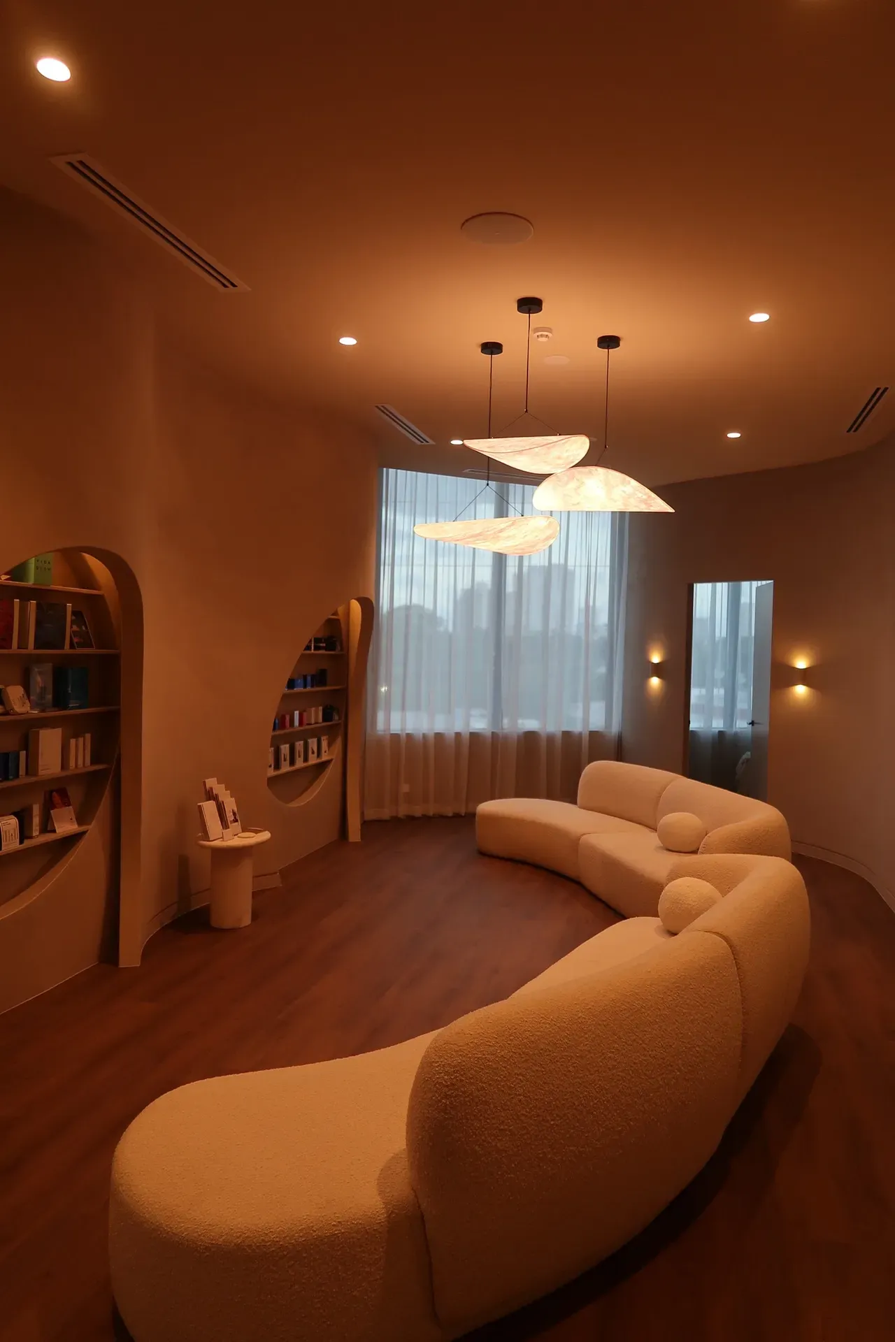 Curved cream sofa in a beige room with a window, shelves, and cloud-shaped pendant lights.