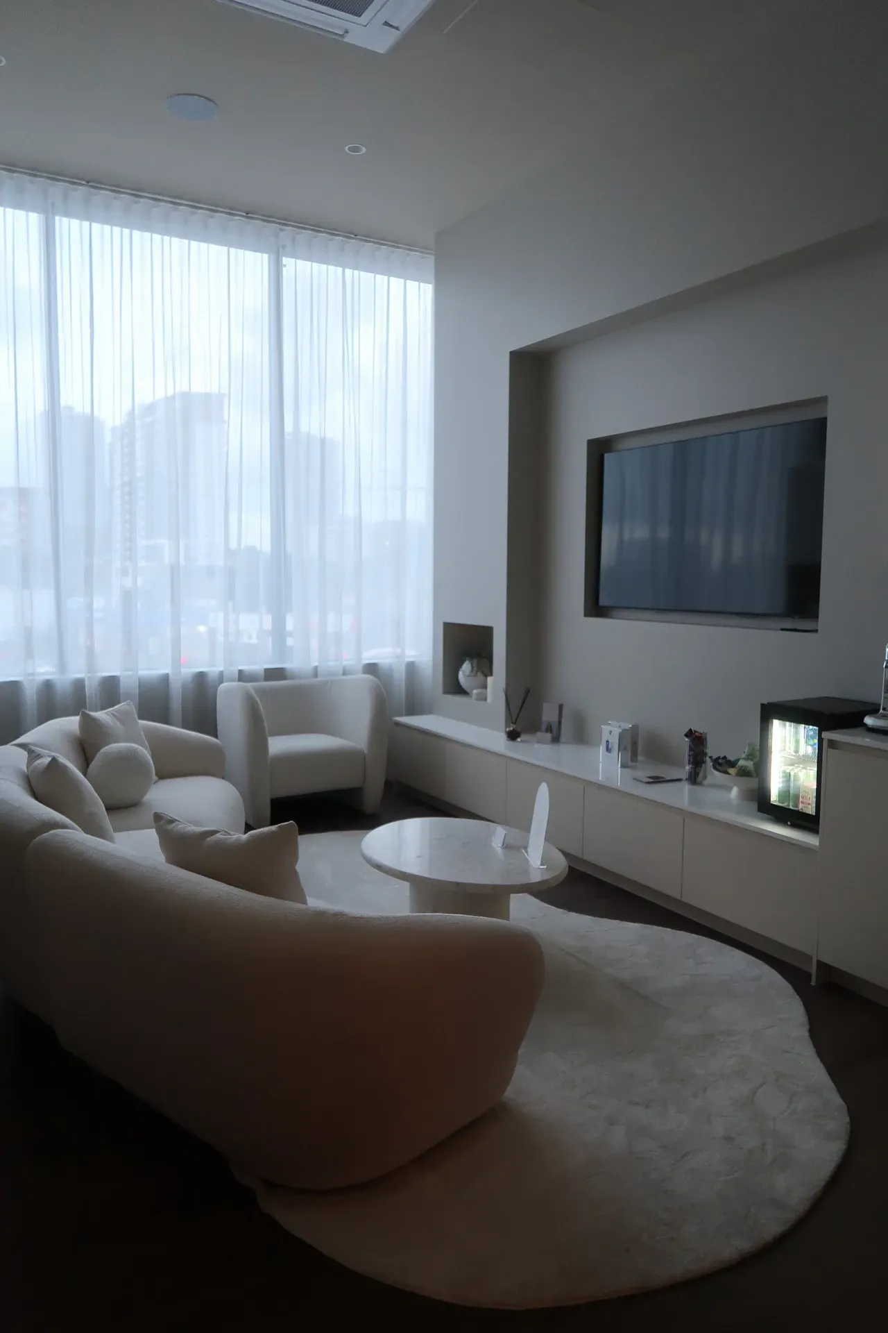 A light-filled, modern living room with cream-colored furniture, TV, and large window with sheer curtains.