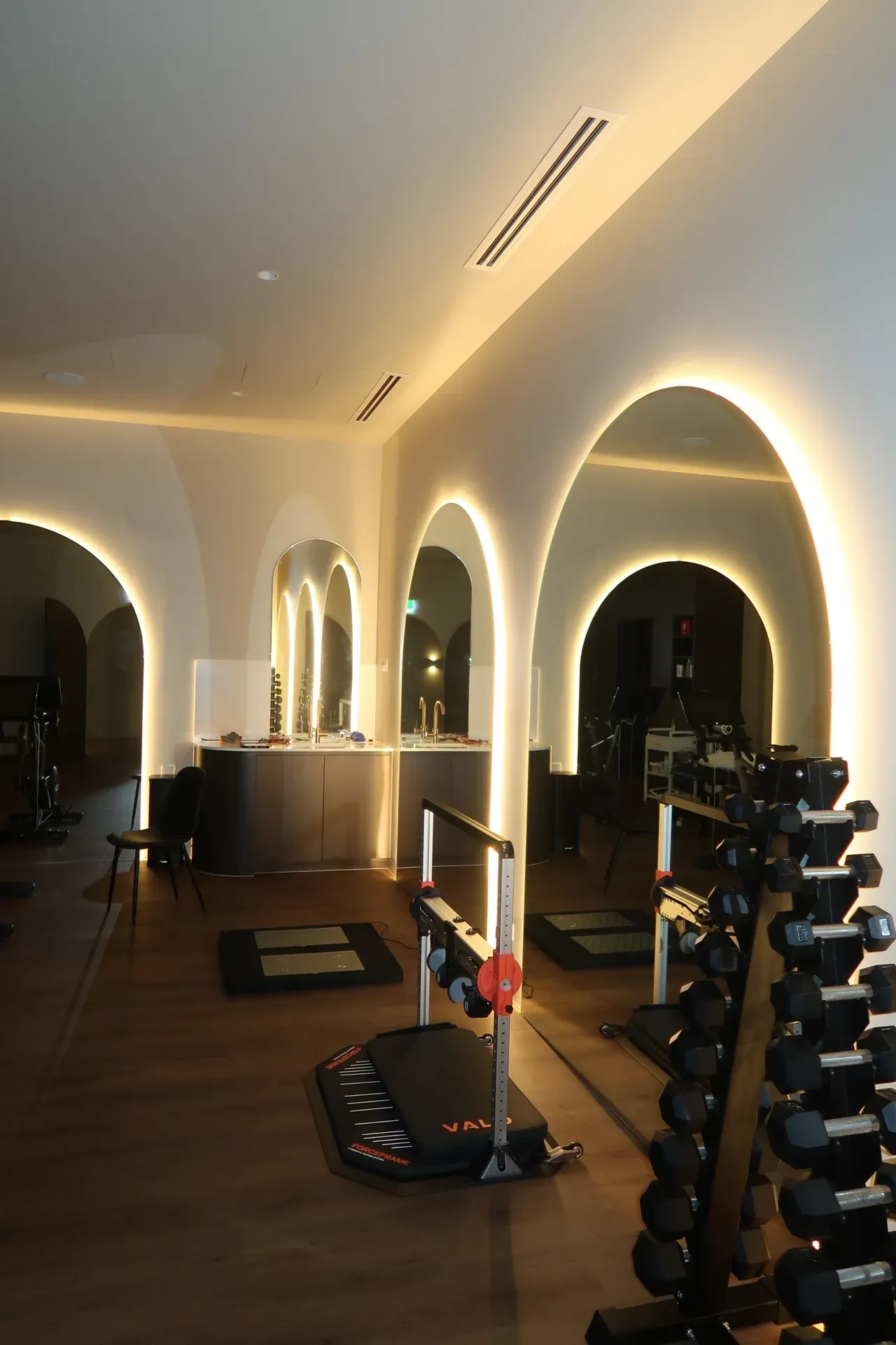 Gym interior with arched mirrors, weights, and workout equipment. Warm lighting. Brown floor, white walls.