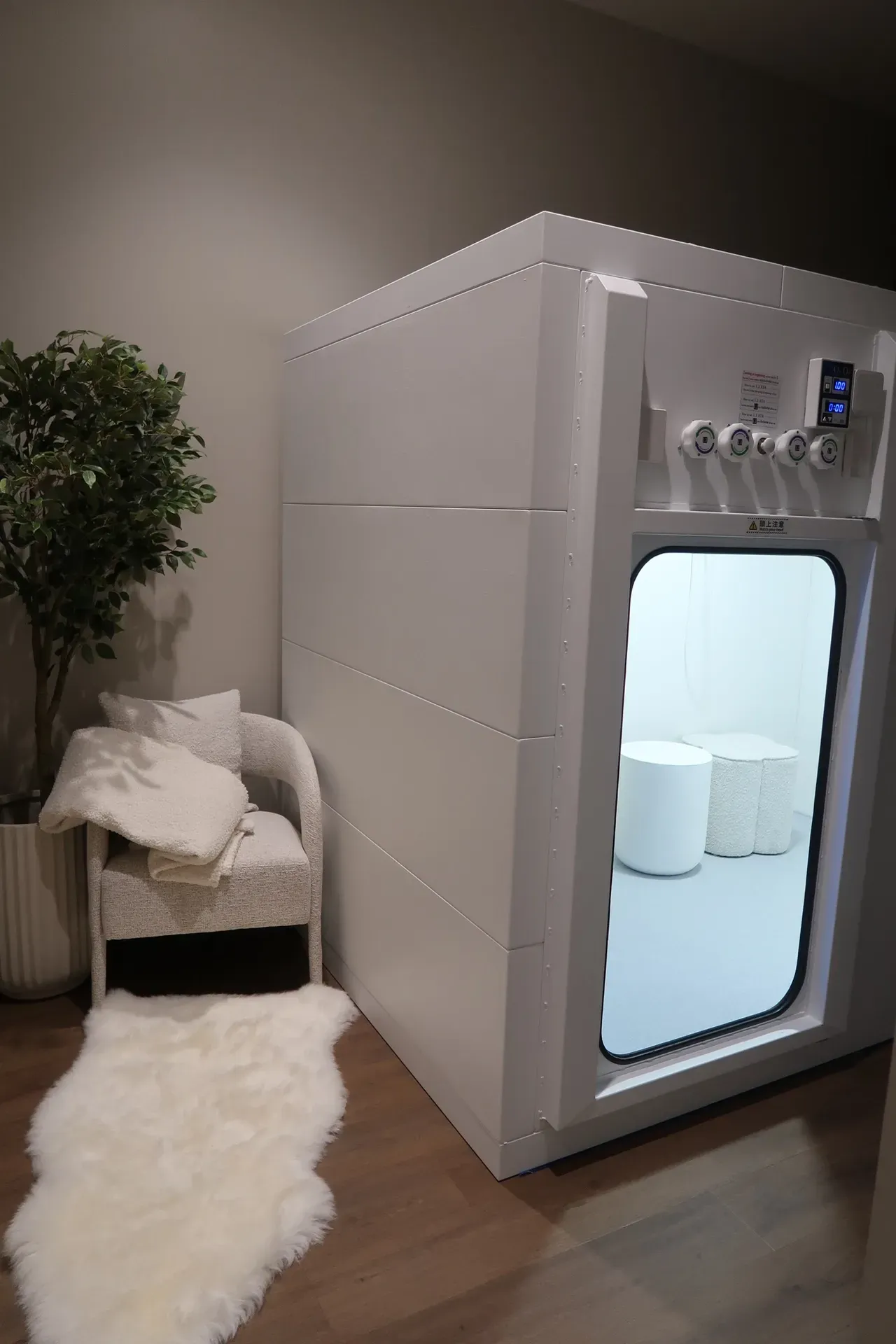 White cryotherapy chamber next to a chair, rug, and plant in a room.