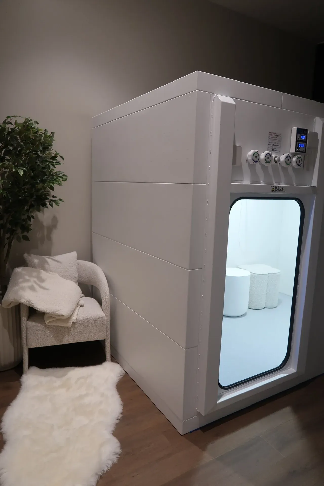 White hyperbaric chamber with window, next to a chair with blanket and a plant.