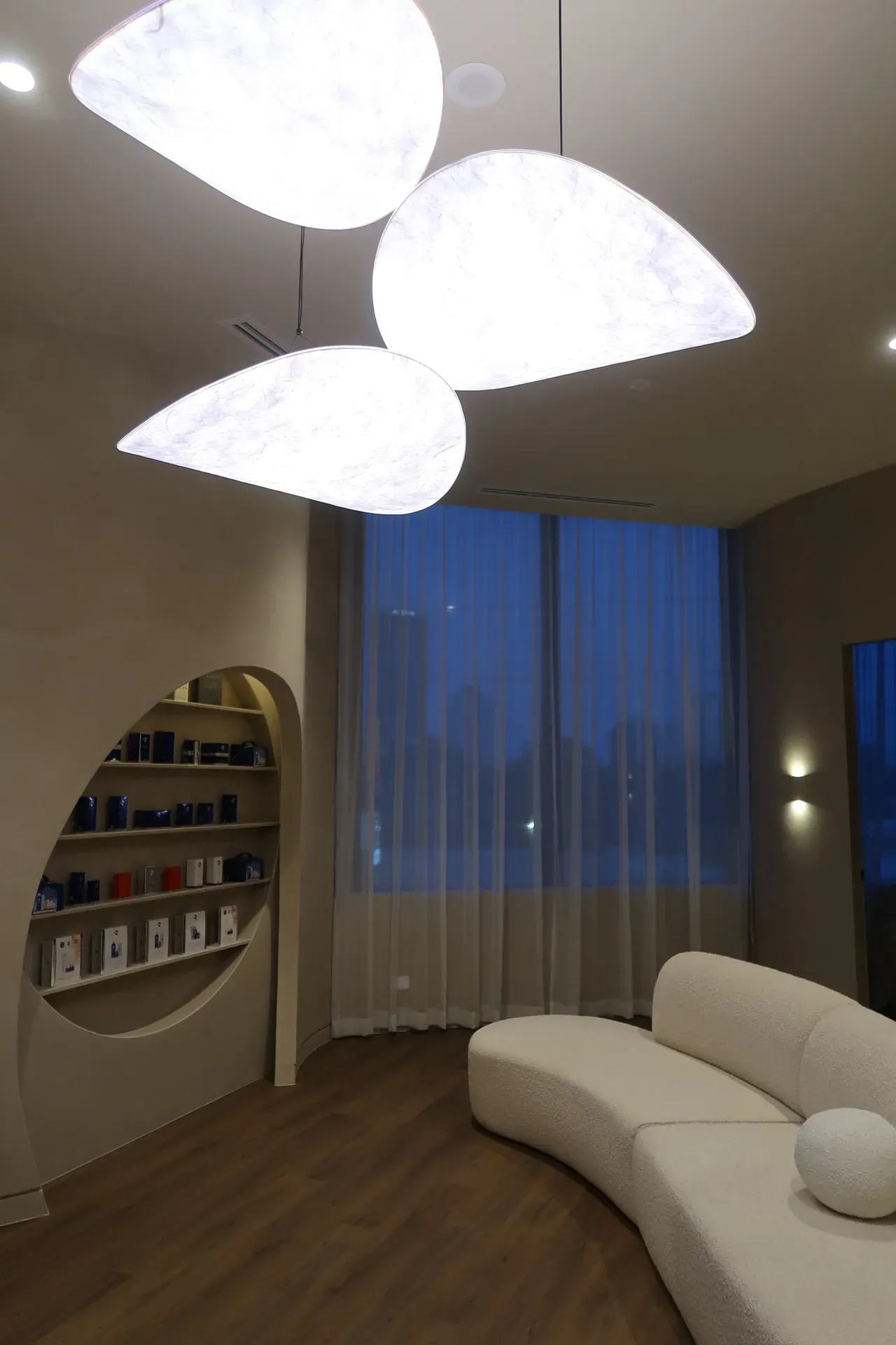 Modern interior with curved white couch, pear-shaped lights, shelves, and window with sheer curtains.