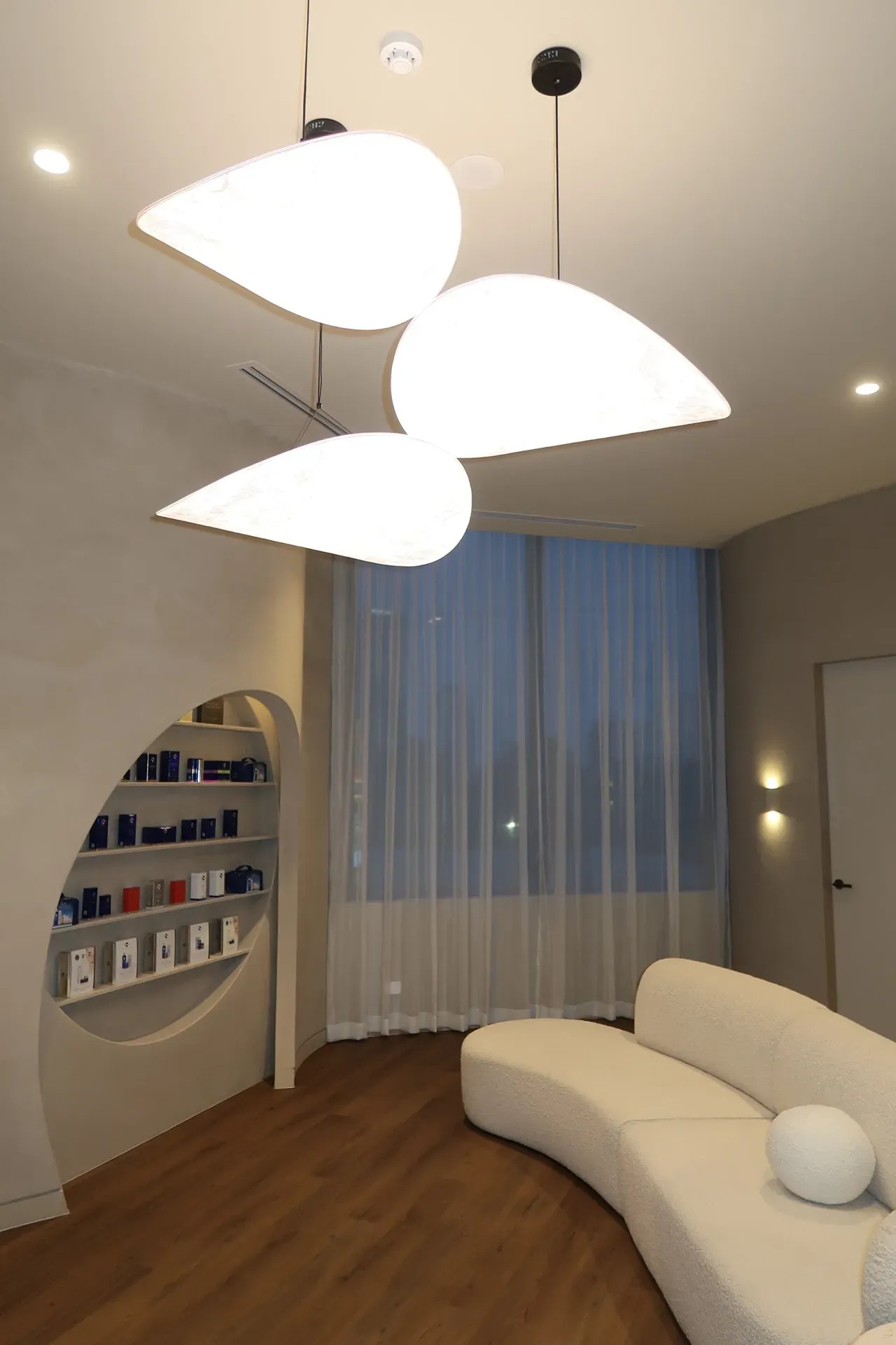 Modern waiting room with curved white sofa, leaf-shaped pendant lights, and built-in product display.