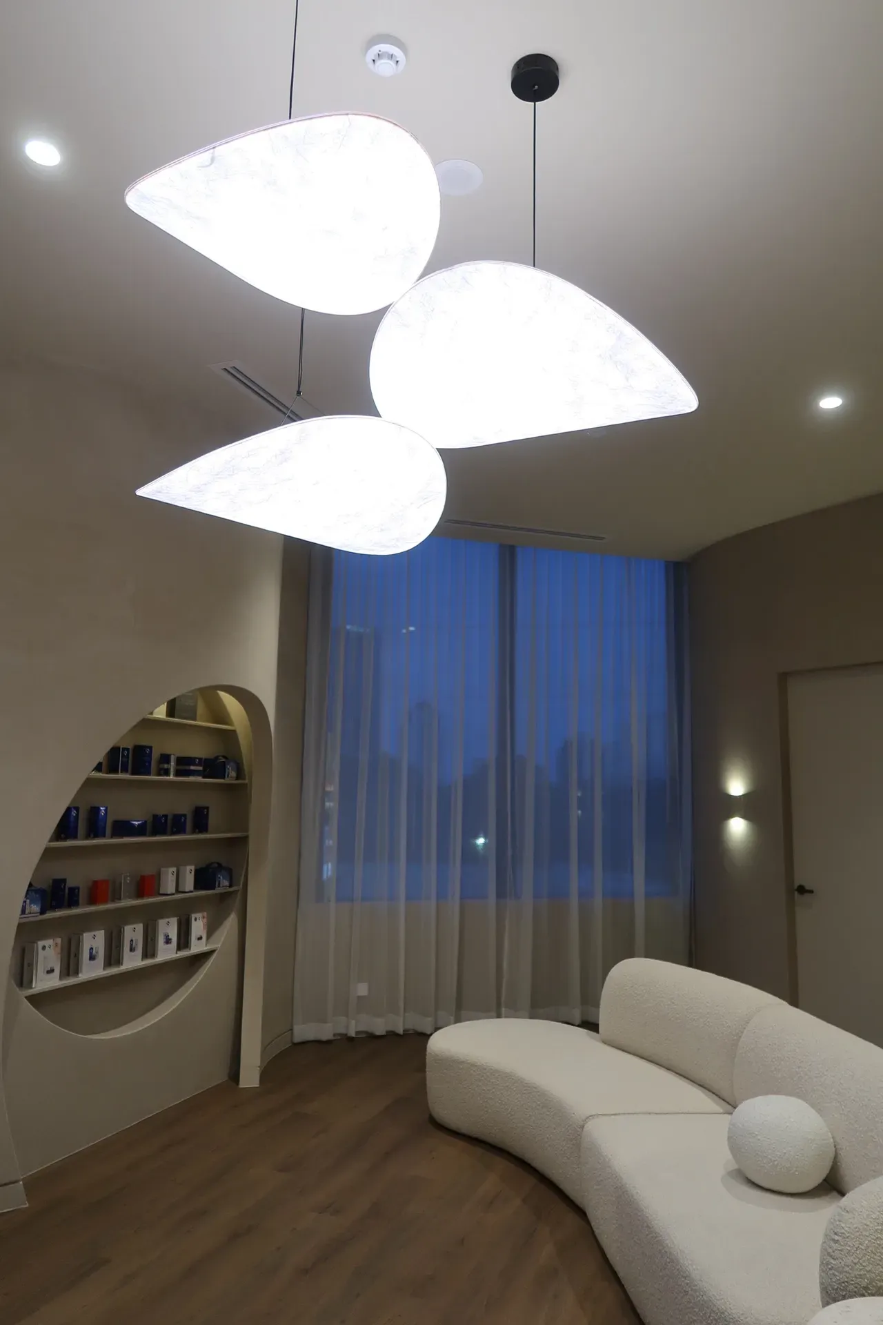 Modern waiting room with curved white sofa, pendant lights, shelving niche, and window with sheer curtains.