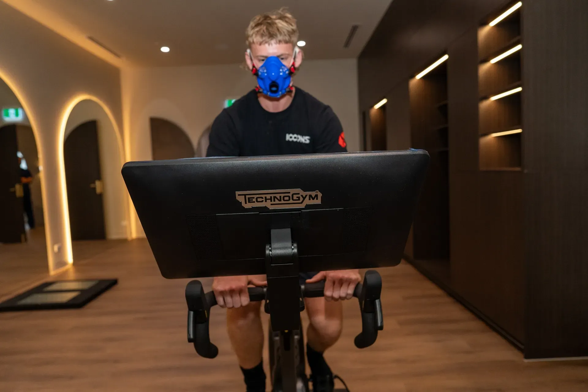 Man wearing a mask, riding a stationary bike, in a modern fitness center.