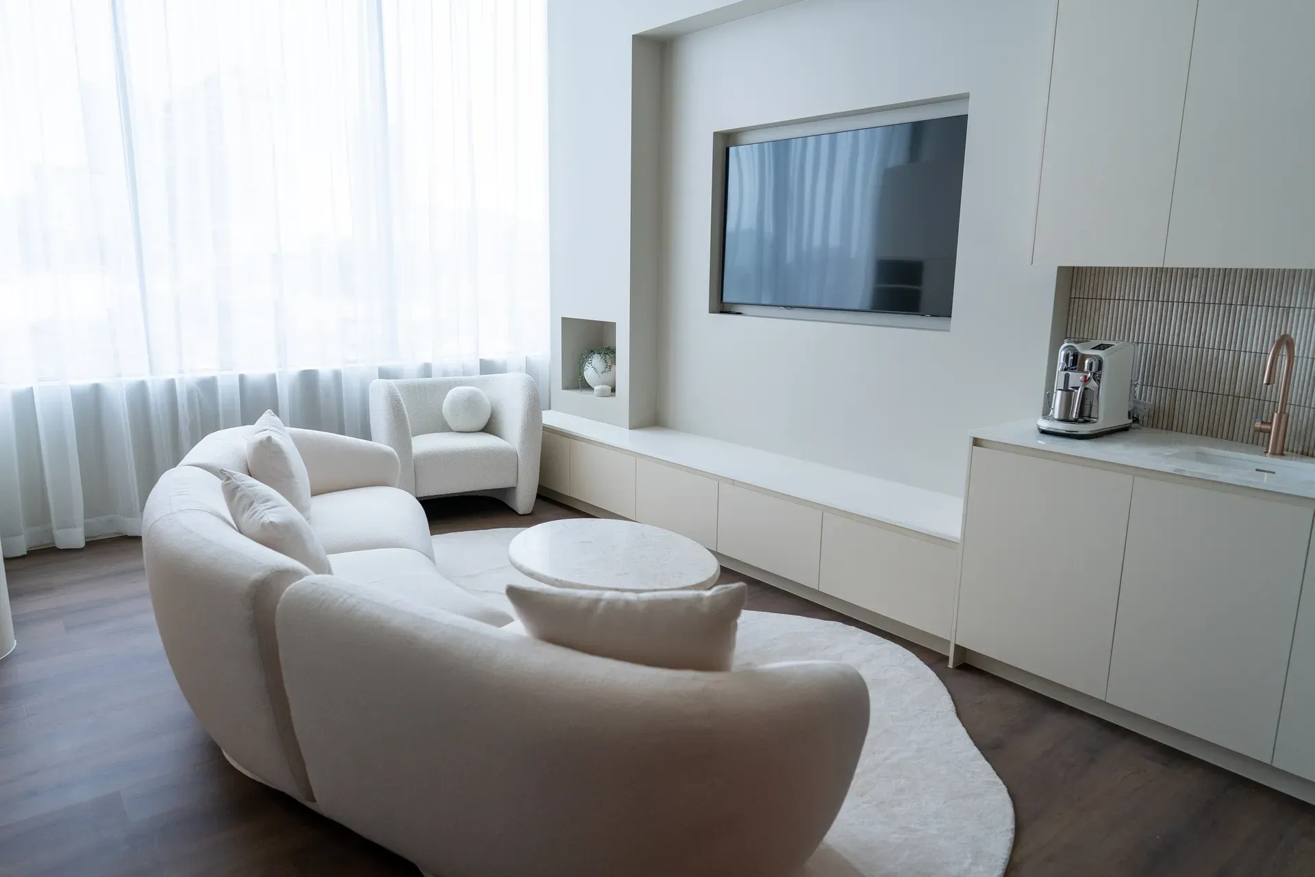 Living room with cream-colored curved sofa, TV, coffee table, and area rug. White walls, window, and cabinetry.