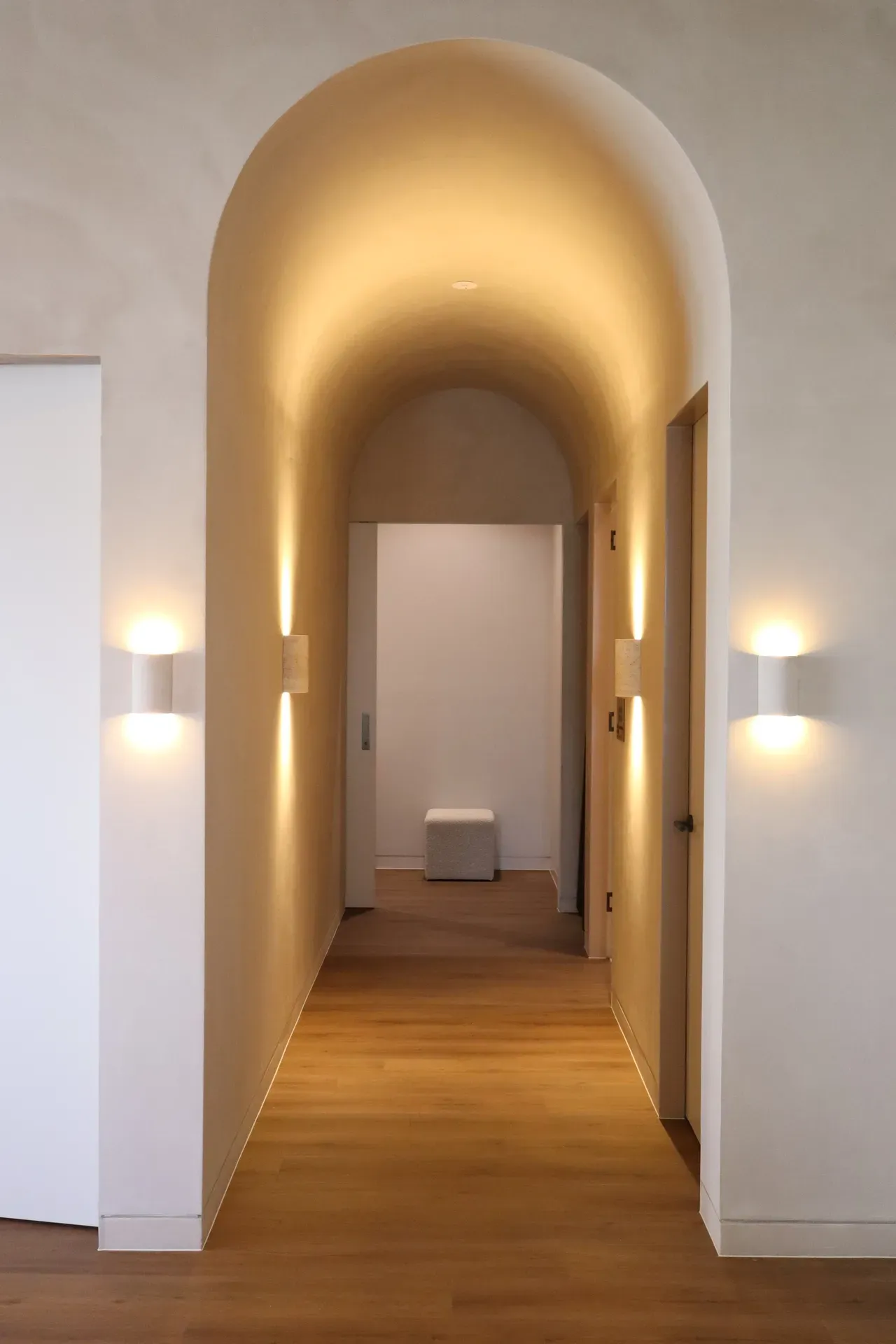 Arched hallway with warm lighting, wooden floor, and modern sconces.