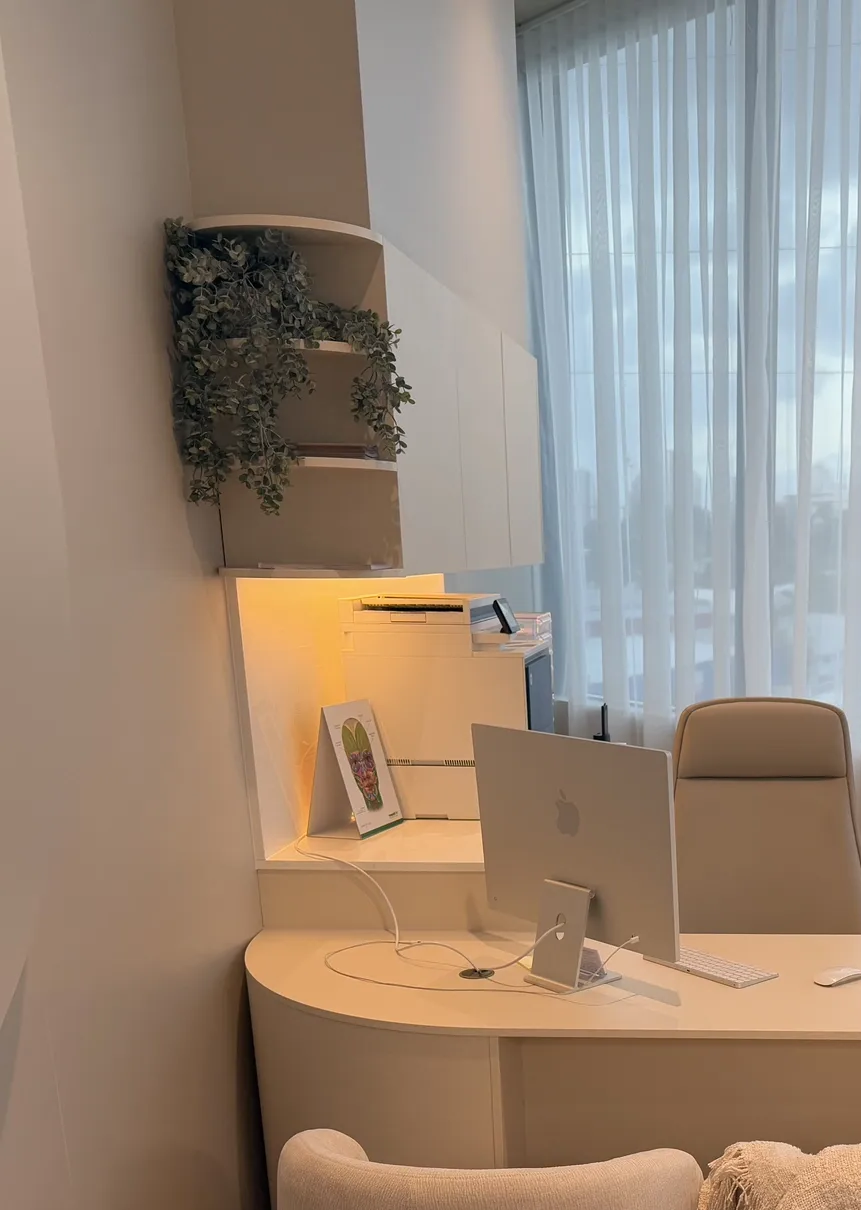 Corner office space with built-in desk, shelves, and a computer. White and beige tones, with a hanging plant and window curtains.