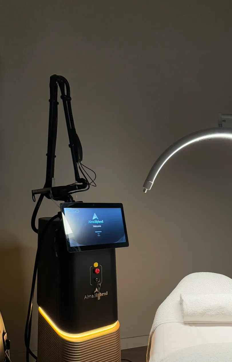 Laser device with touch screen, next to curved light and treatment table in a dimly lit room.