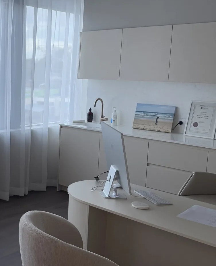 Modern office with beige desk, computer, and chair. White cabinets, sink, and artwork by a window with sheer curtains.