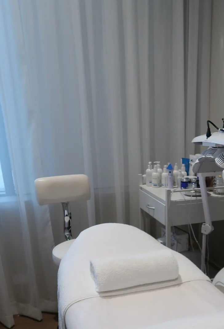 Treatment room: white bed with rolled towel, chair, table with products, white curtains.