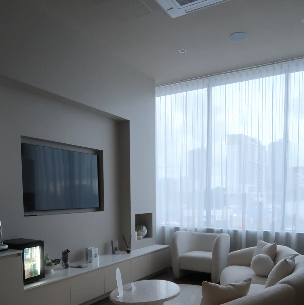A modern living room with a TV, a sofa, and large windows with sheer curtains.