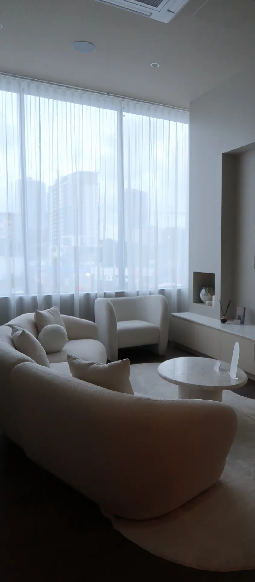 A modern, light-filled living room with white furniture, sheer curtains, and a round coffee table.