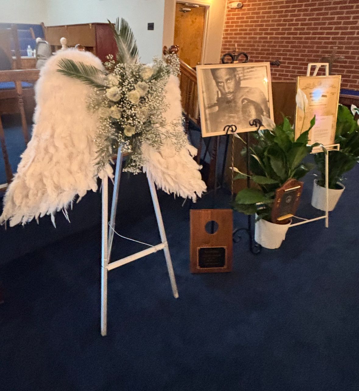 Mourning display: floral angel wings, framed photo, plants, and wooden plaque on blue carpet.