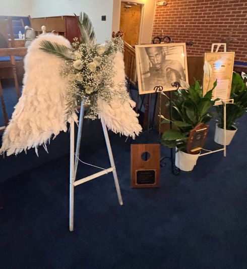 Mourning display: floral angel wings, framed photo, plants, and wooden plaque on blue carpet.