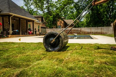 A lawn roller moves over a patch of grass in a backyard with a house and pool in the background.