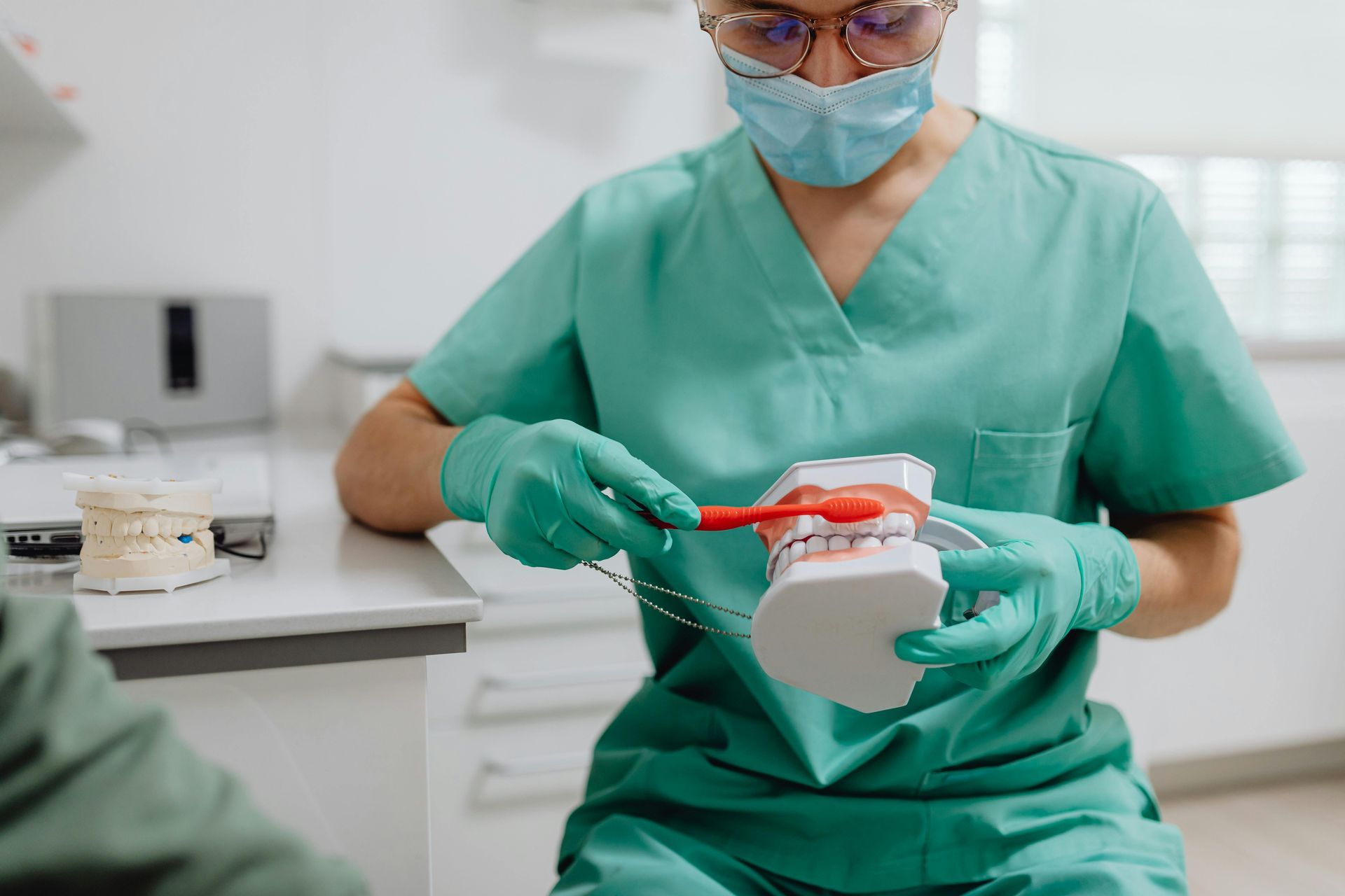 A dental professional in green scrubs and a mask uses a red toothbrush to demonstrate proper cleaning on a tooth model.