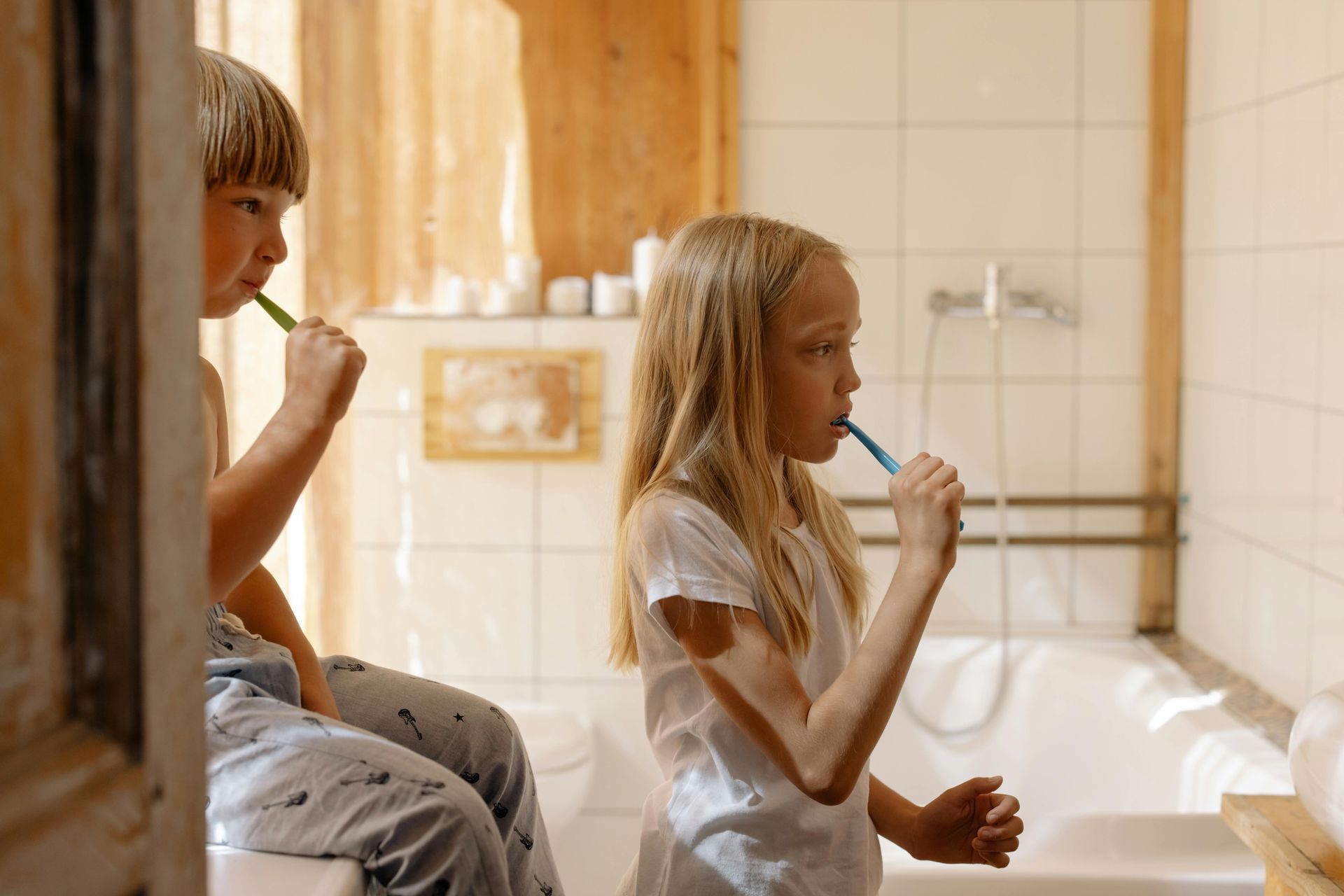 Two children in white shirts brush their teeth in a tiled bathroom with a bathtub.