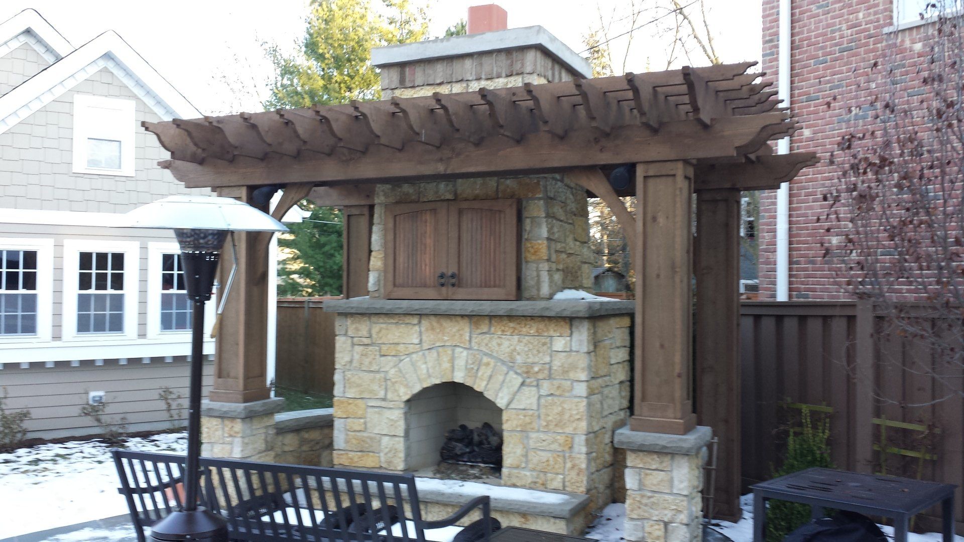 Outdoor fireplace with stone facade, wooden pergola, and snow on the ground.