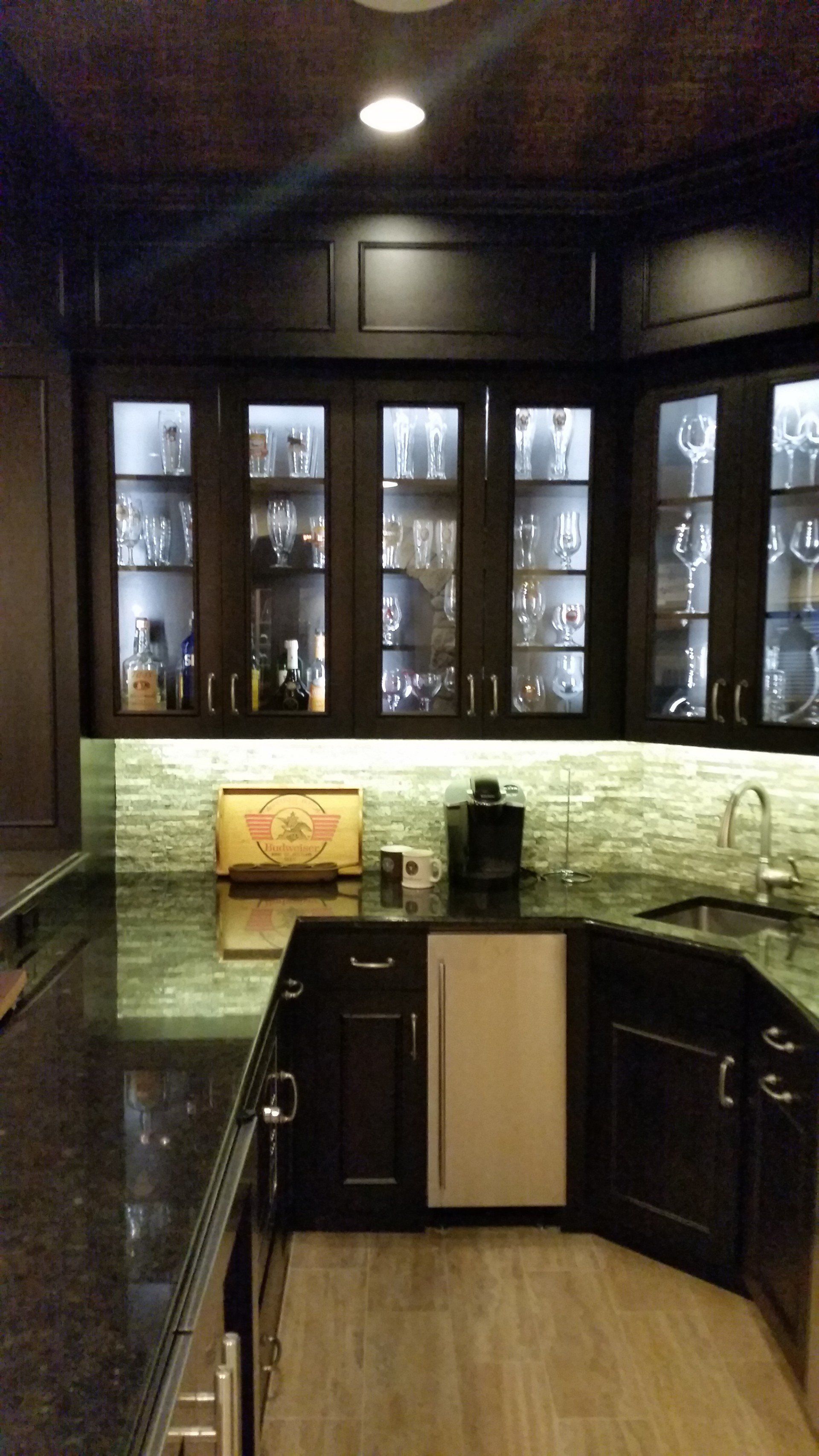 Dark wood home bar with glass cabinets, a small refrigerator, and a green tile backsplash.