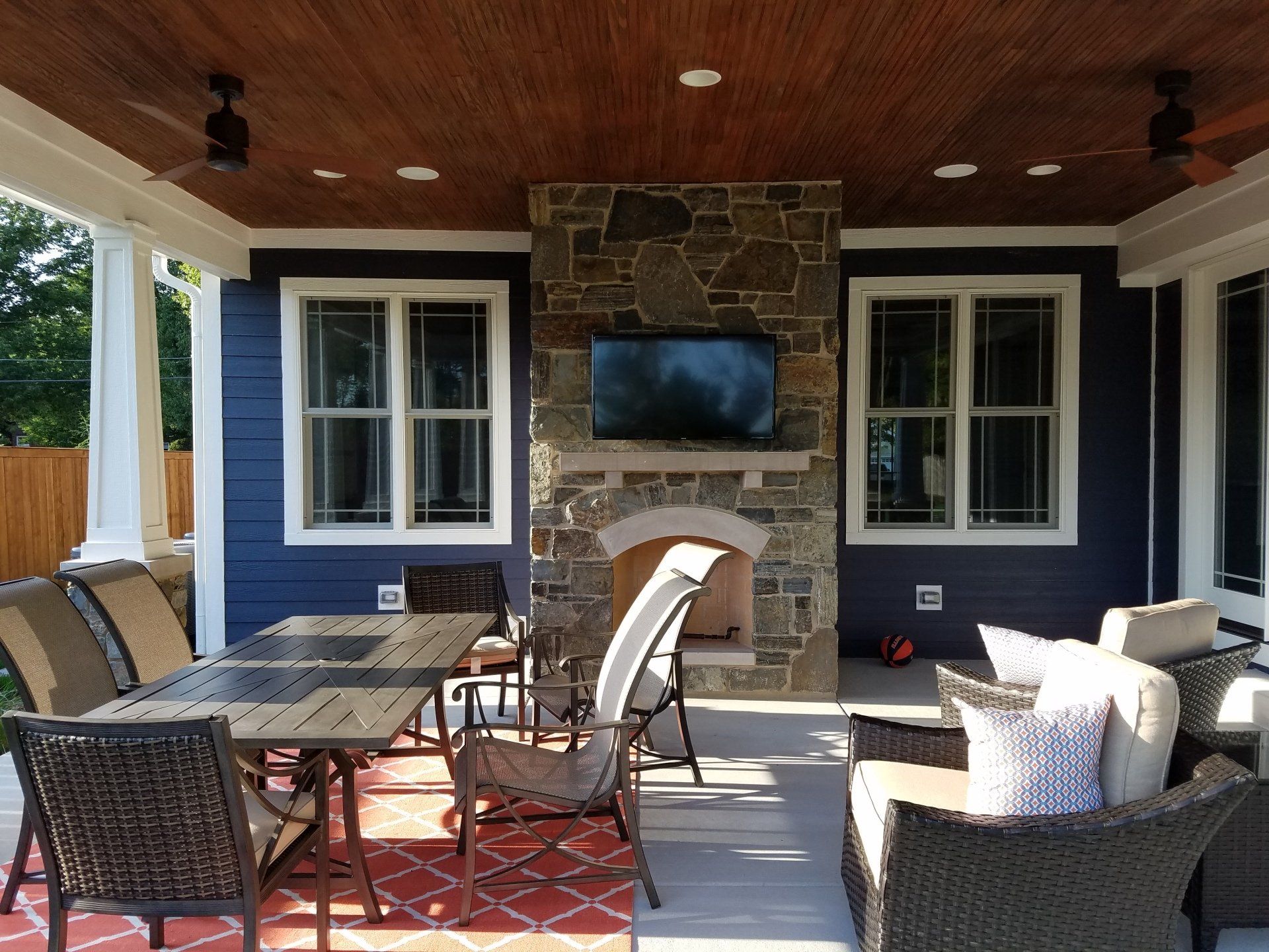 Outdoor patio with fireplace, seating, table, and TV. Blue siding, stone fireplace, wood ceiling.
