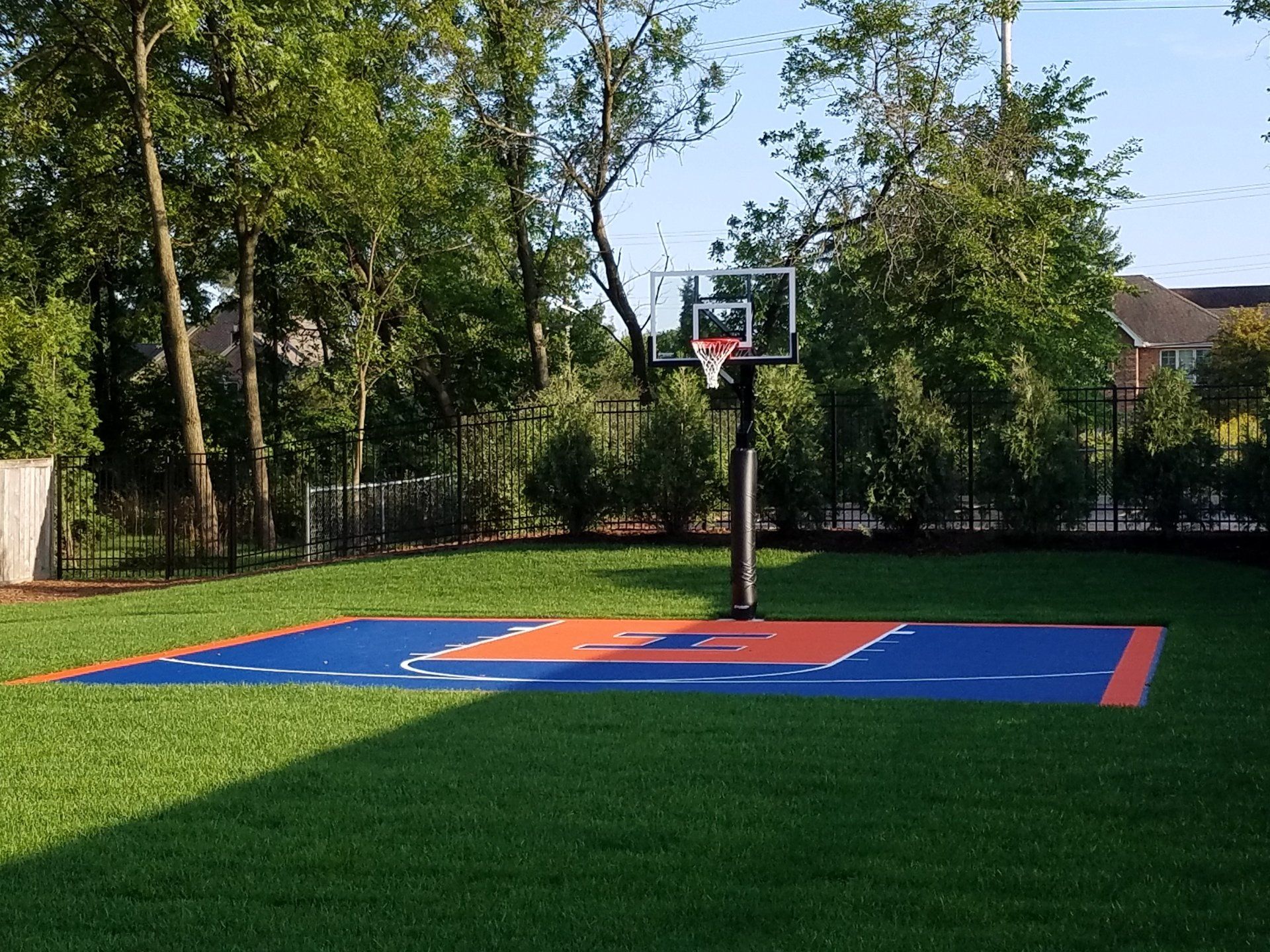 Basketball hoop on blue and orange court, set on a grassy lawn with trees in the background.