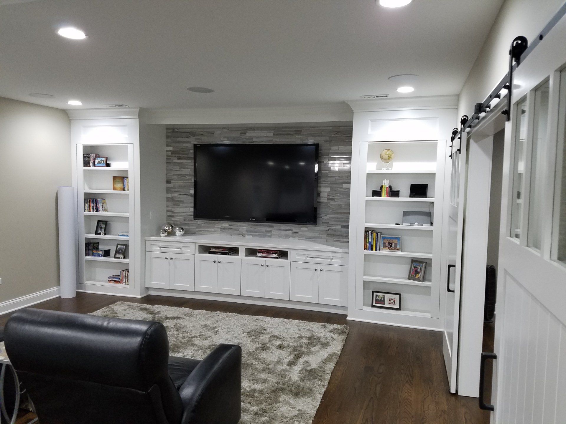 Living room with built-in white cabinets, large TV on stone wall, black leather chair, and a rug.