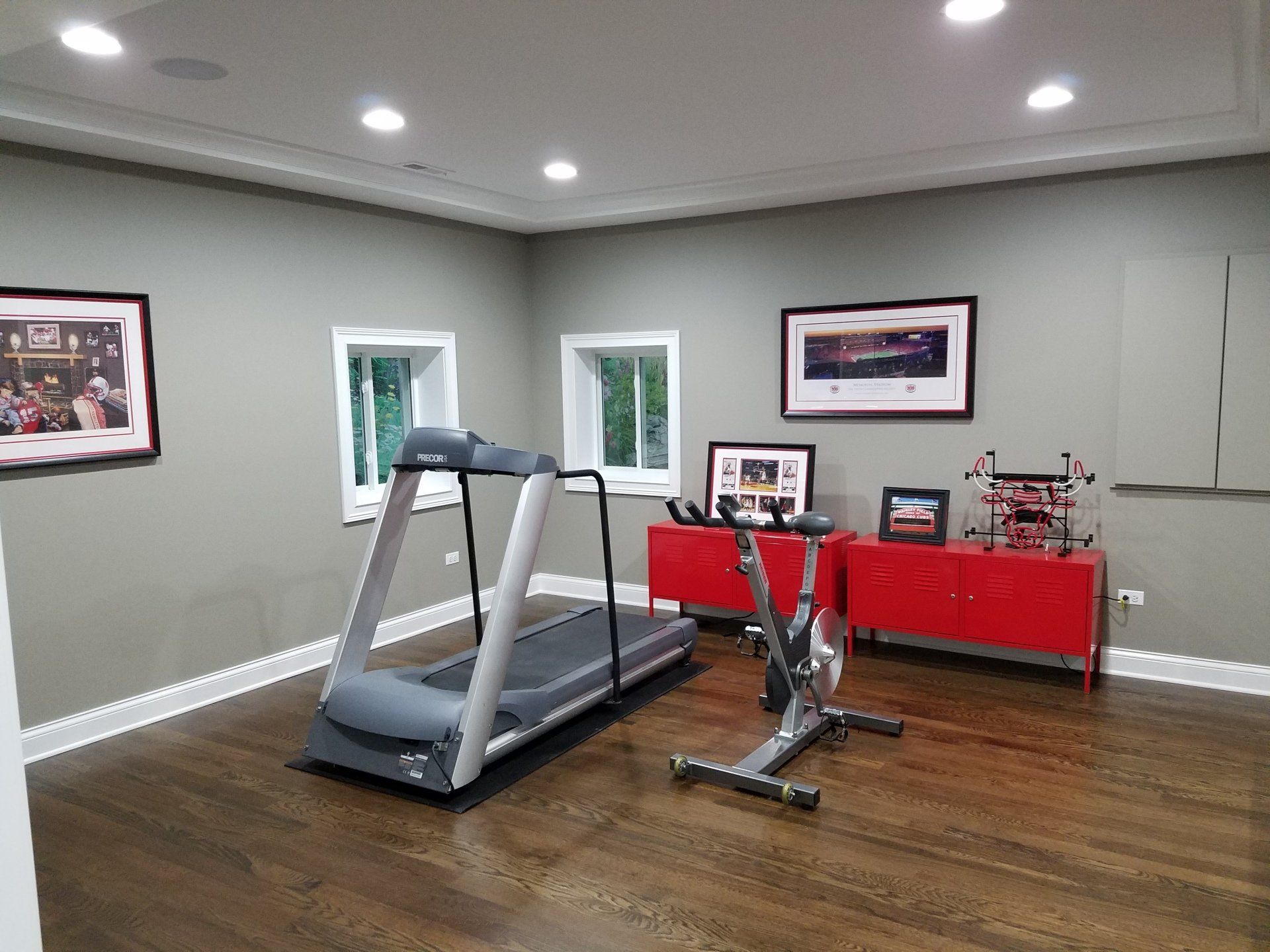 Home gym with a treadmill, exercise bike, and red storage cabinets against gray walls and wood floor.
