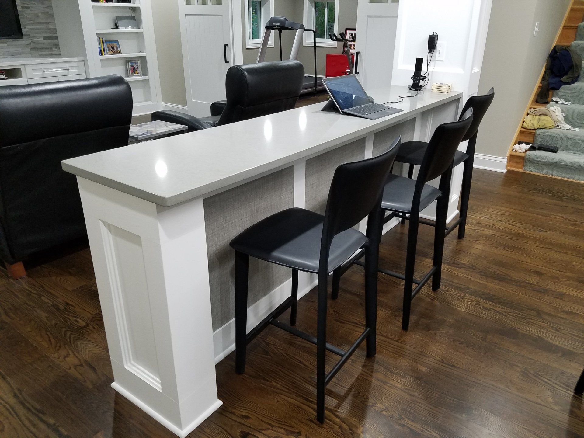 A long, white bar with gray countertop, black barstools, in a home basement with dark wood flooring.